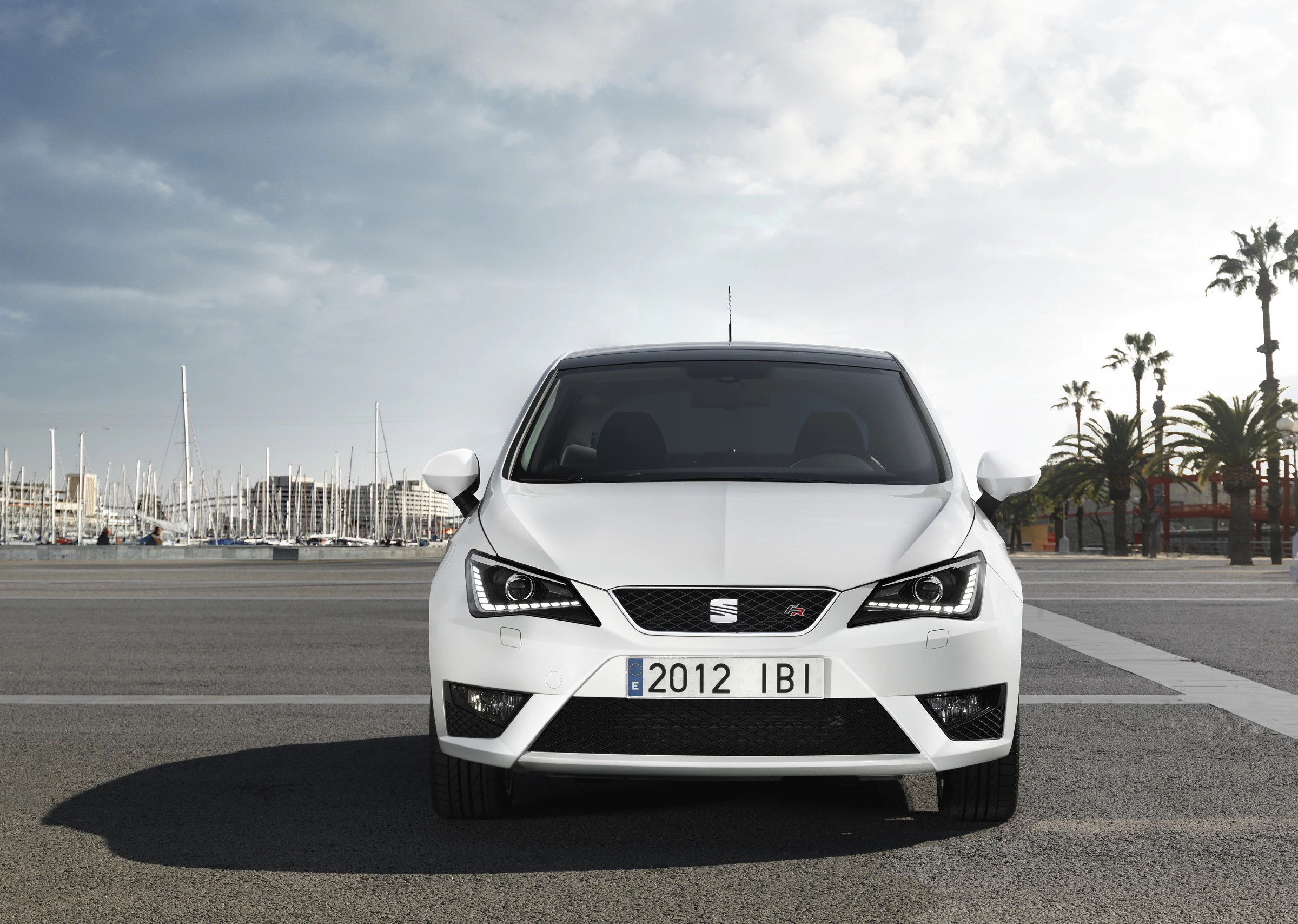 Weißer SEAT Ibiza FR steht auf einem Parkplatz mit Hafenhintergrund; Palmen und Boote sind im Hintergrund zu sehen.