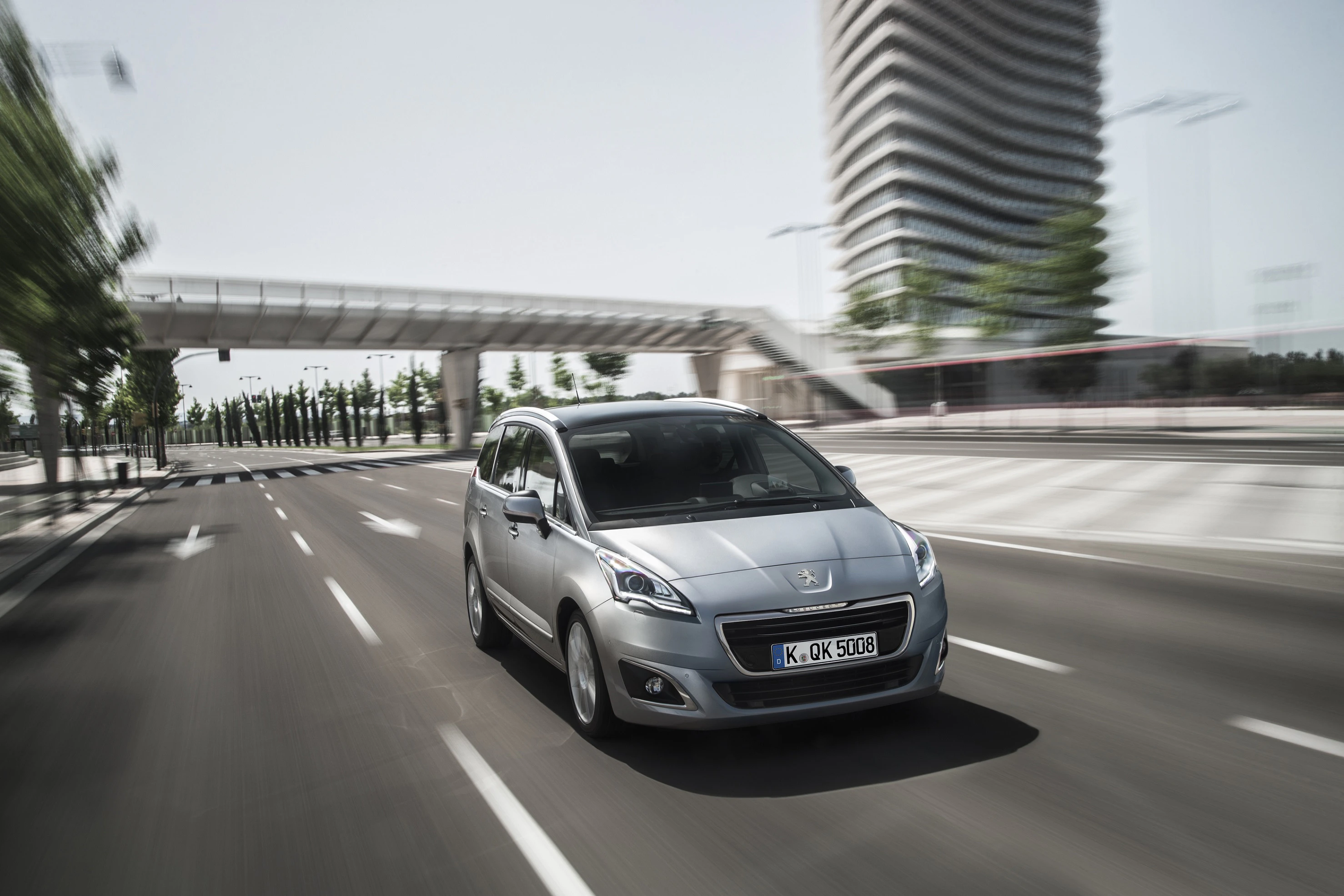 Silberner Peugeot 5008 fährt auf leerer Straße mit moderner Architektur im Hintergrund. Dynamische Bewegungsunschärfe.