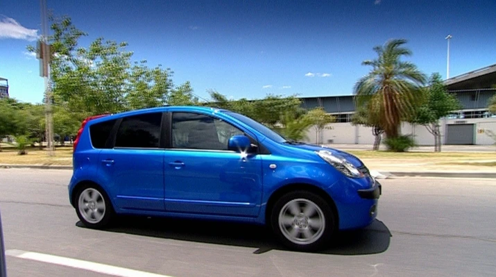 Blauer Kleinwagen fährt auf einer Straße, mit Palmen und Gebäuden im Hintergrund bei klarem Himmel.