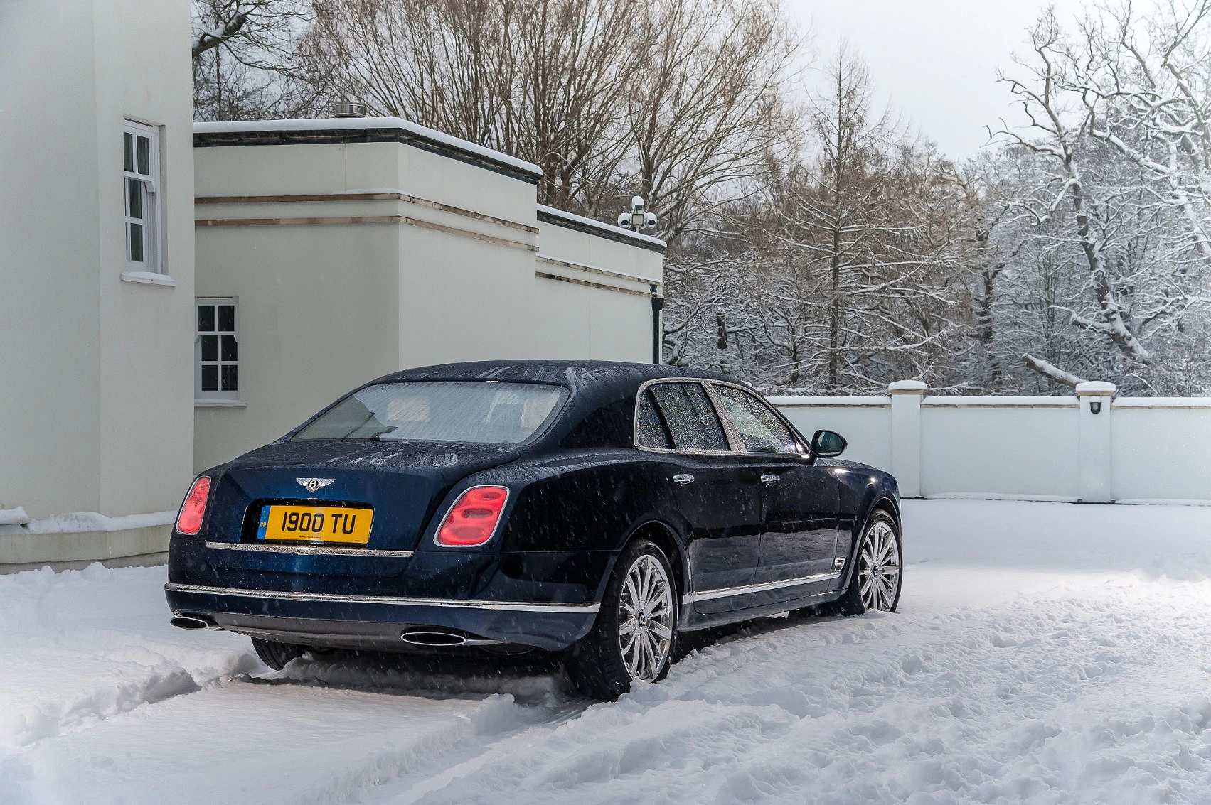 Ein blauer Luxuswagen parkt im verschneiten Hof eines Gebäudes. Schneebedeckte Bäume sind im Hintergrund sichtbar.