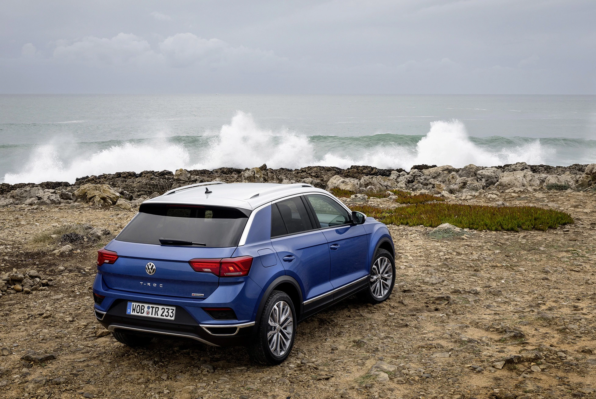 Blauer SUV auf felsigem Küstenweg mit Blick auf stürmische Meeresbrandung unter bewölktem Himmel.