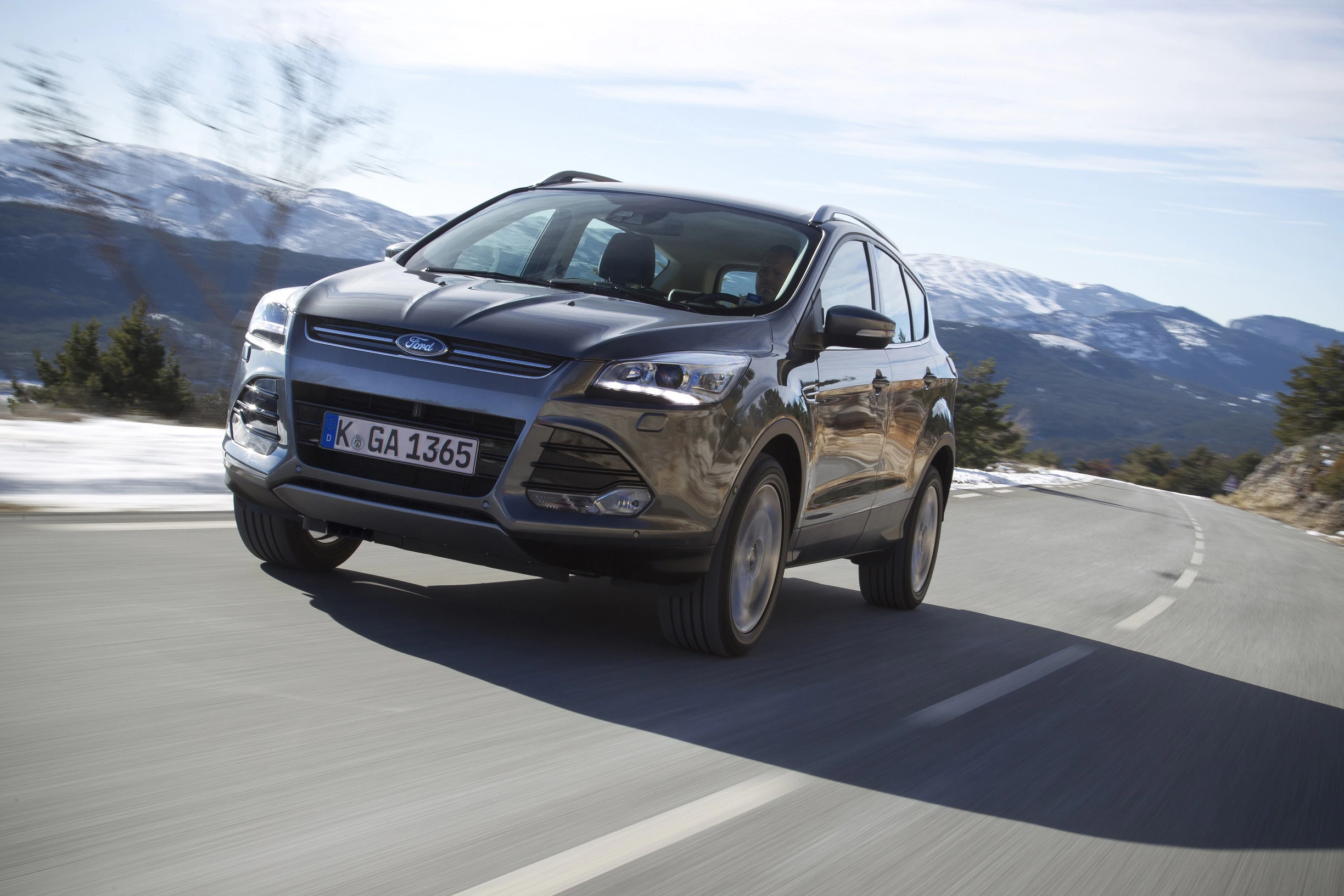 Ein grauer SUV fährt auf einer Landstraße in einer bergigen, verschneiten Landschaft unter bewölktem Himmel.