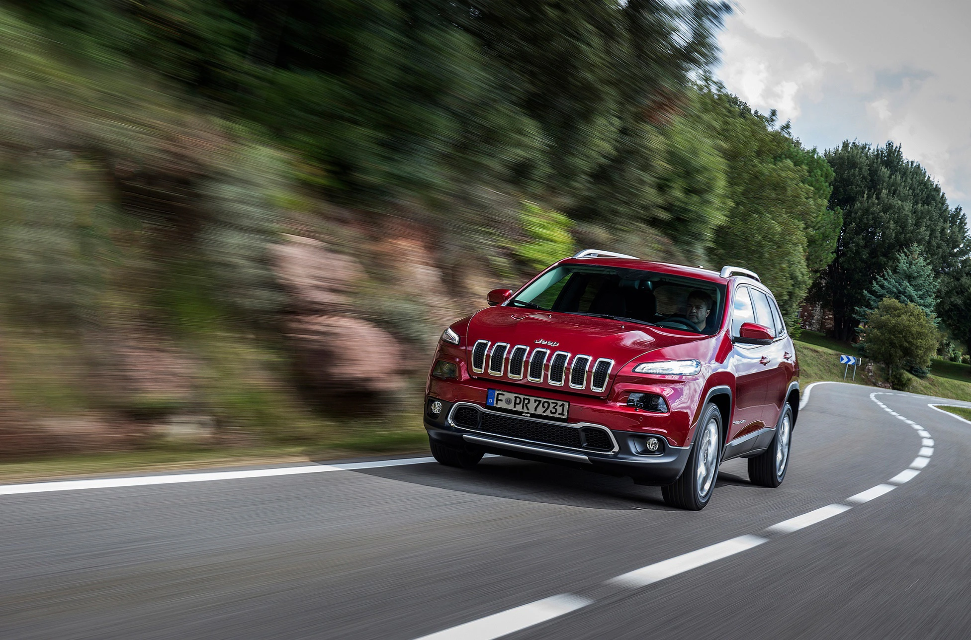 Roter SUV fährt auf einer kurvigen Landstraße durch eine grüne Landschaft, bei bewölktem Himmel und leichter Bewegungsunschärfe.
