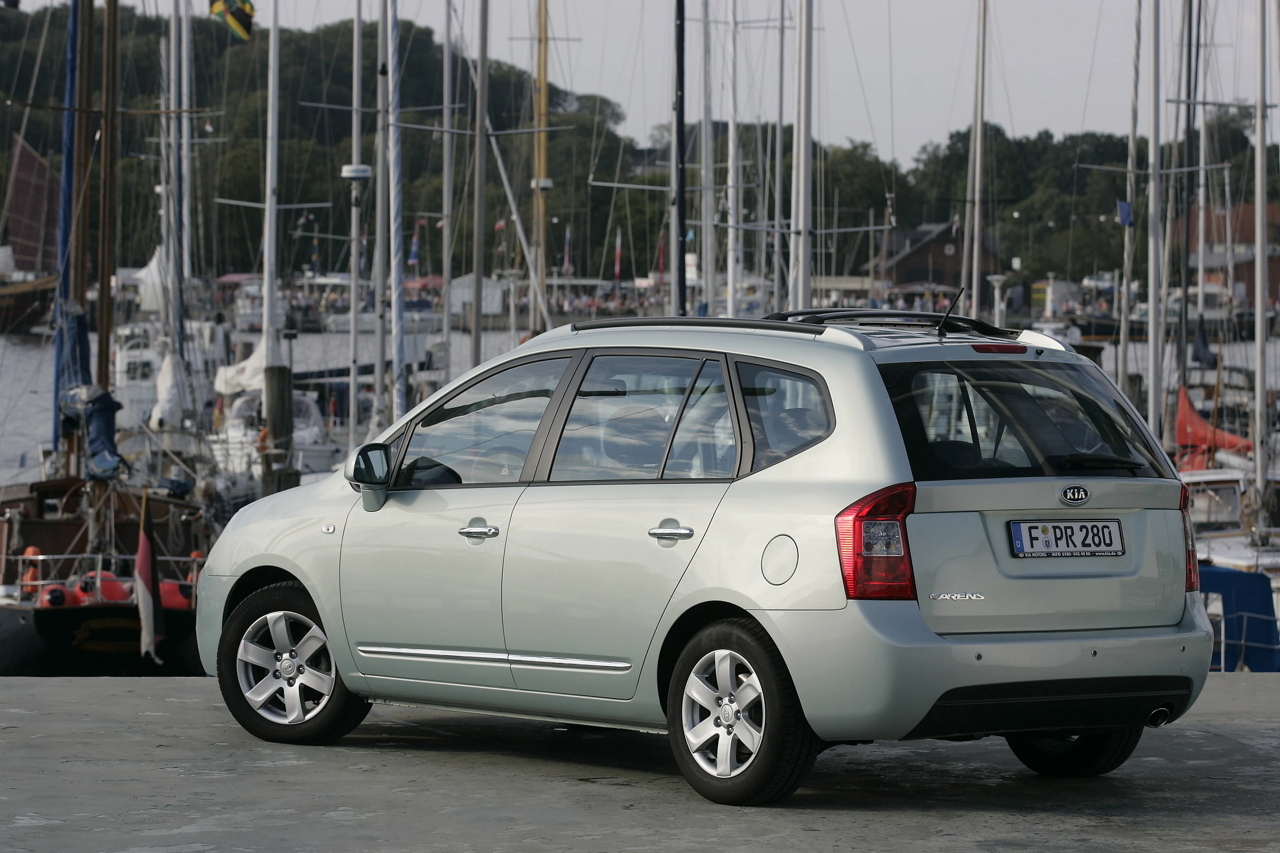 Ein silberner Minivan parkt an einem Yachthafen, umgeben von Segelbooten und Masten. Im Hintergrund sind Bäume sichtbar.