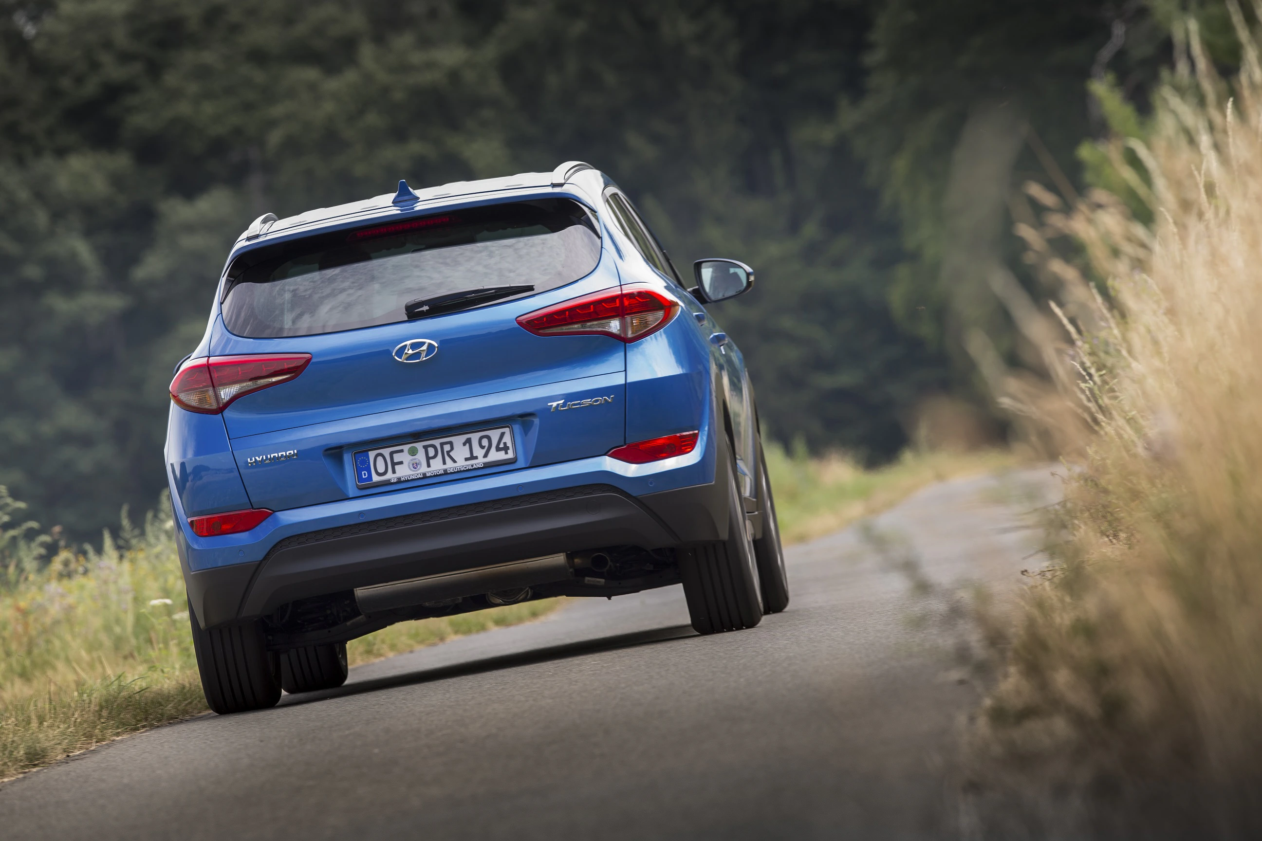 Blauer Hyundai Tucson auf schmaler Landstraße, umgeben von Bäumen und Gras am Straßenrand.