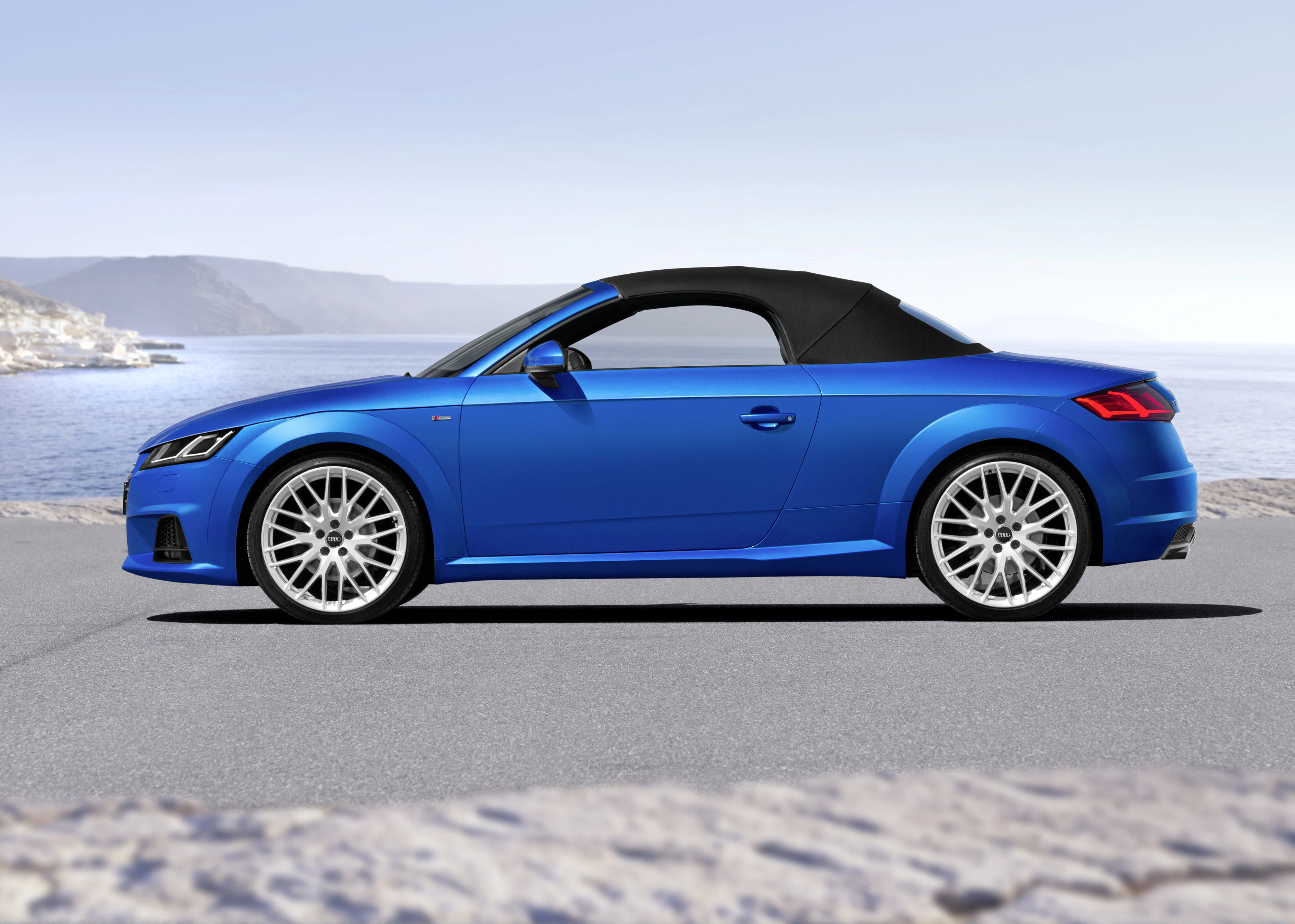 Blauer Sportwagen-Cabrio auf Küstenstraße, Seitenansicht, mit schwarzem Stoffverdeck und silbernen Felgen. Meer und Hügel im Hintergrund.