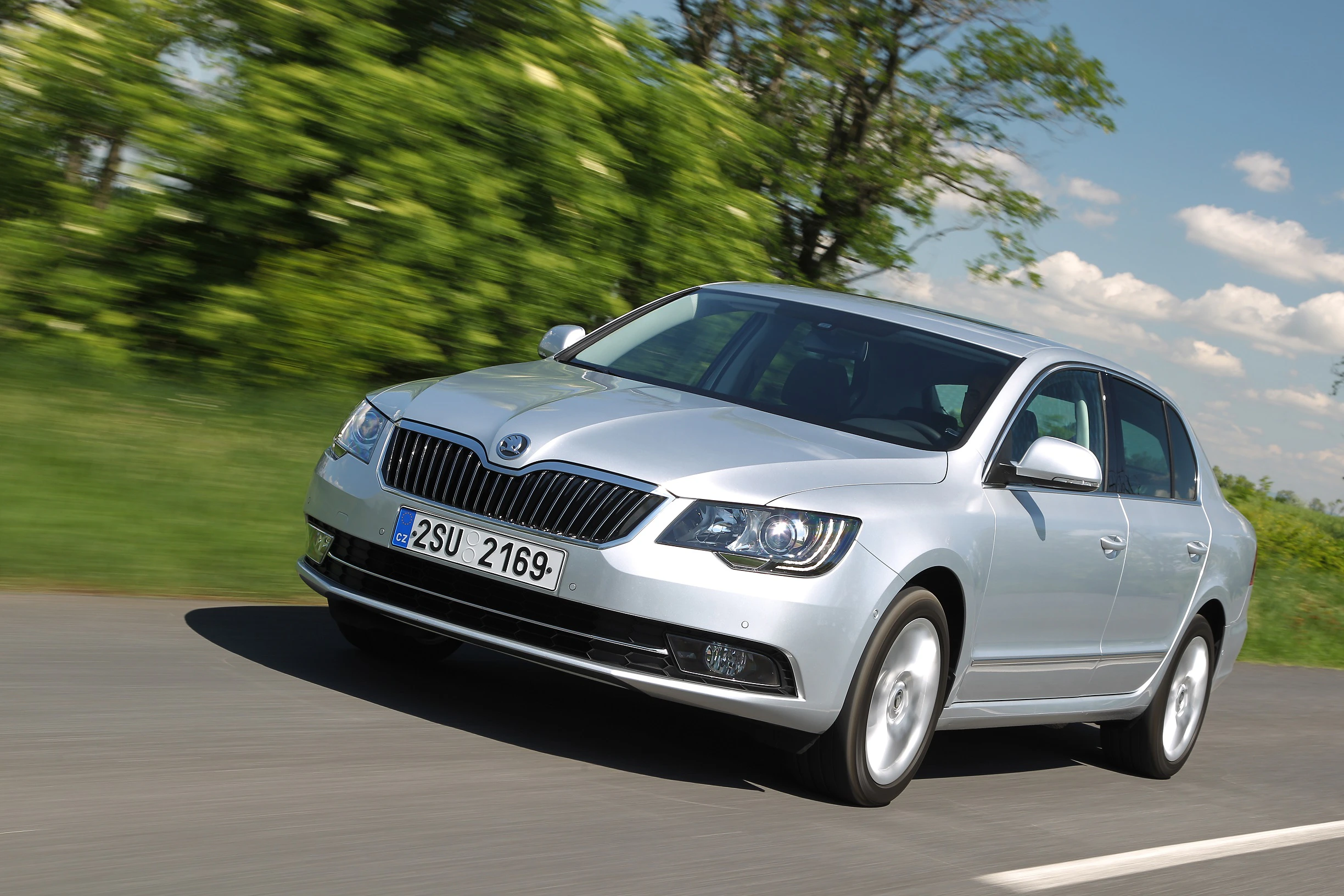 Silberner Skoda fährt schnell auf einer Landstraße, umgeben von grünen Bäumen und blauem Himmel.