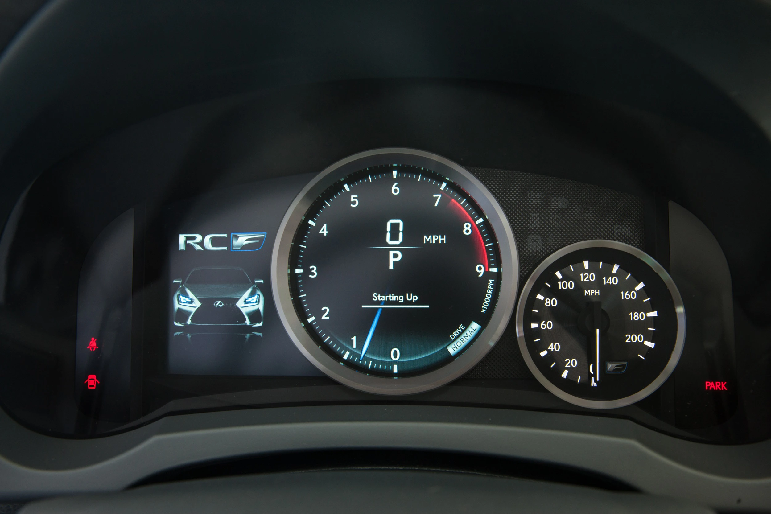 Nahaufnahme eines Fahrzeugarmaturenbretts mit digitalem Tacho, Drehzahlmesser und einem Display, das ein Auto und "Starting Up" zeigt.