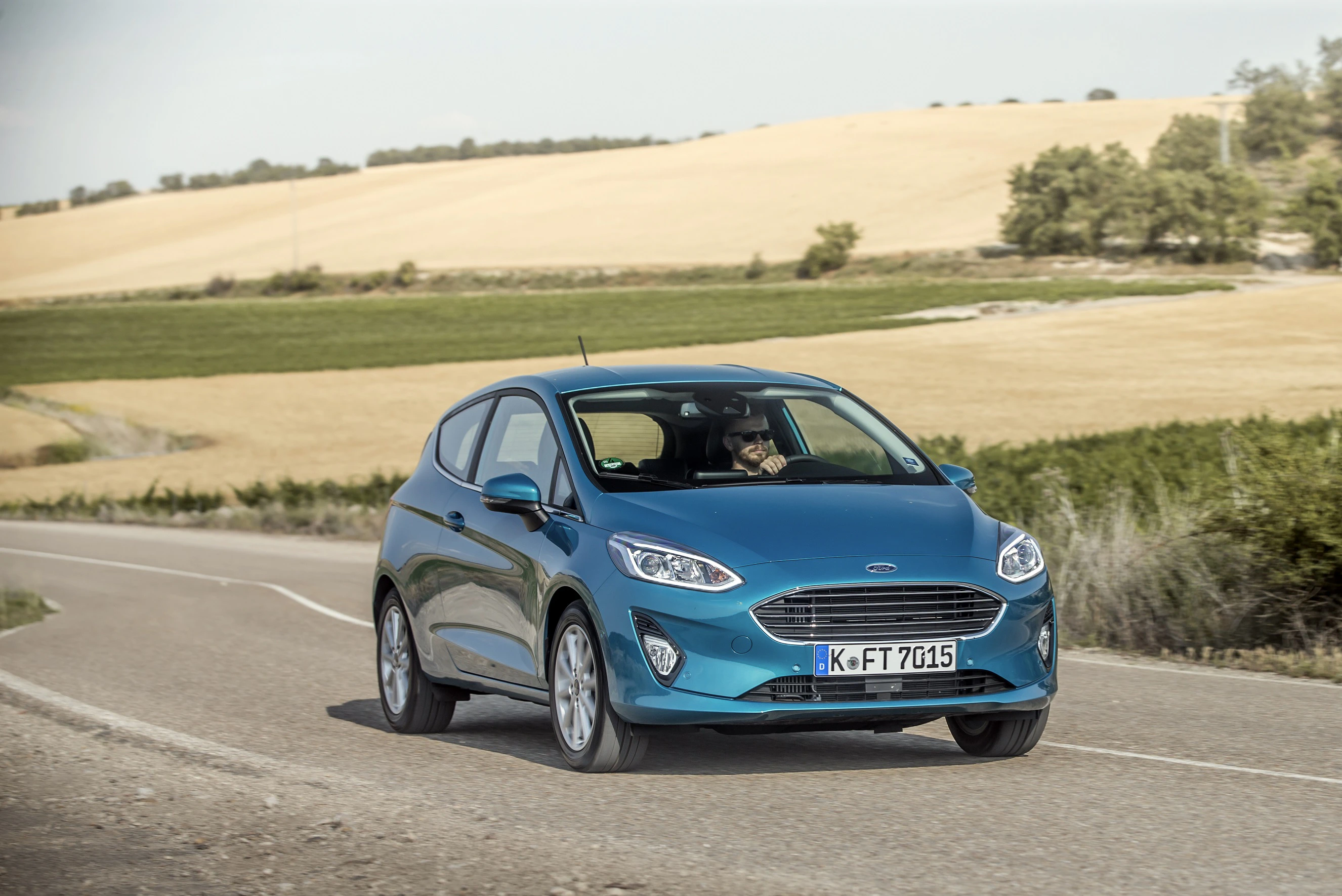 Blauer Kleinwagen auf einer Landstraße, umgeben von grünen Feldern und sanften Hügeln unter blauem Himmel.