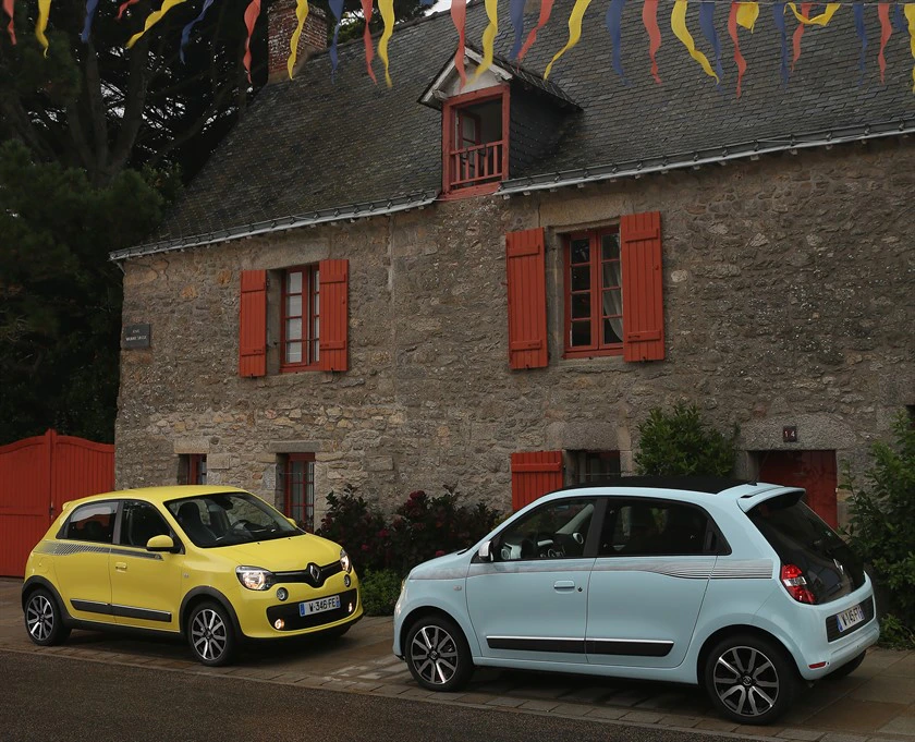 Zwei kleine Autos, eines gelb und eines hellblau, parken vor einem alten Steinhaus mit roten Fensterläden und Girlanden darüber.