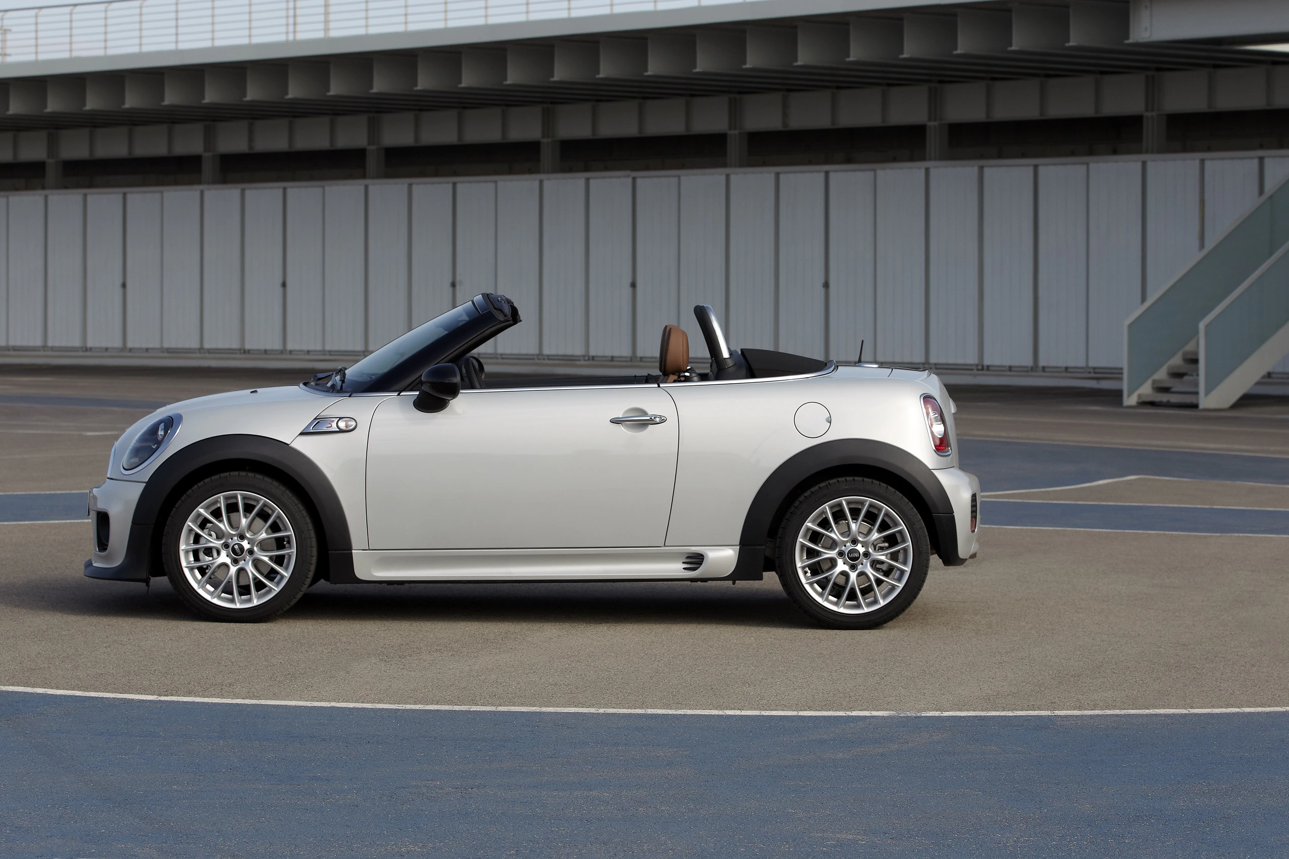 Silbernes Cabrio in einem leeren Areal, mit offenem Verdeck und deutlich sichtbaren Reifen, seitliche Ansicht.
