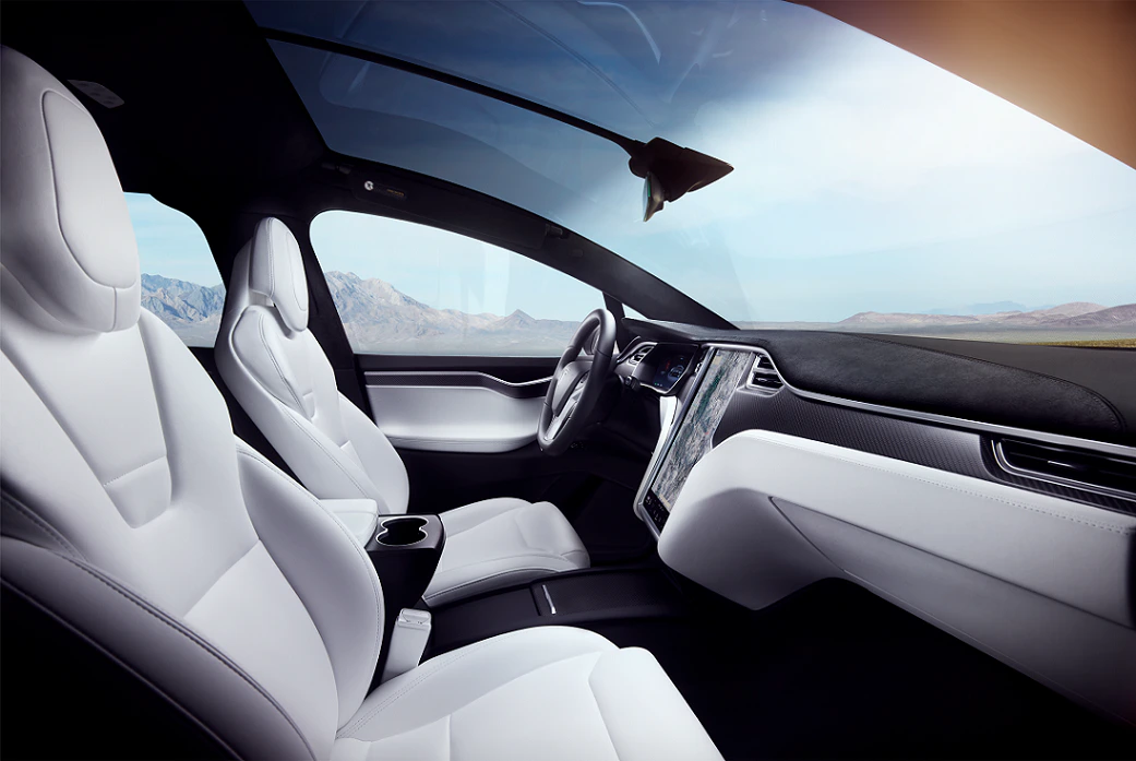 Innenraum eines modernen Autos mit weißen Ledersitzen, digitalem Display und Panorama-Glasdach, mit Blick auf eine bergige Landschaft.