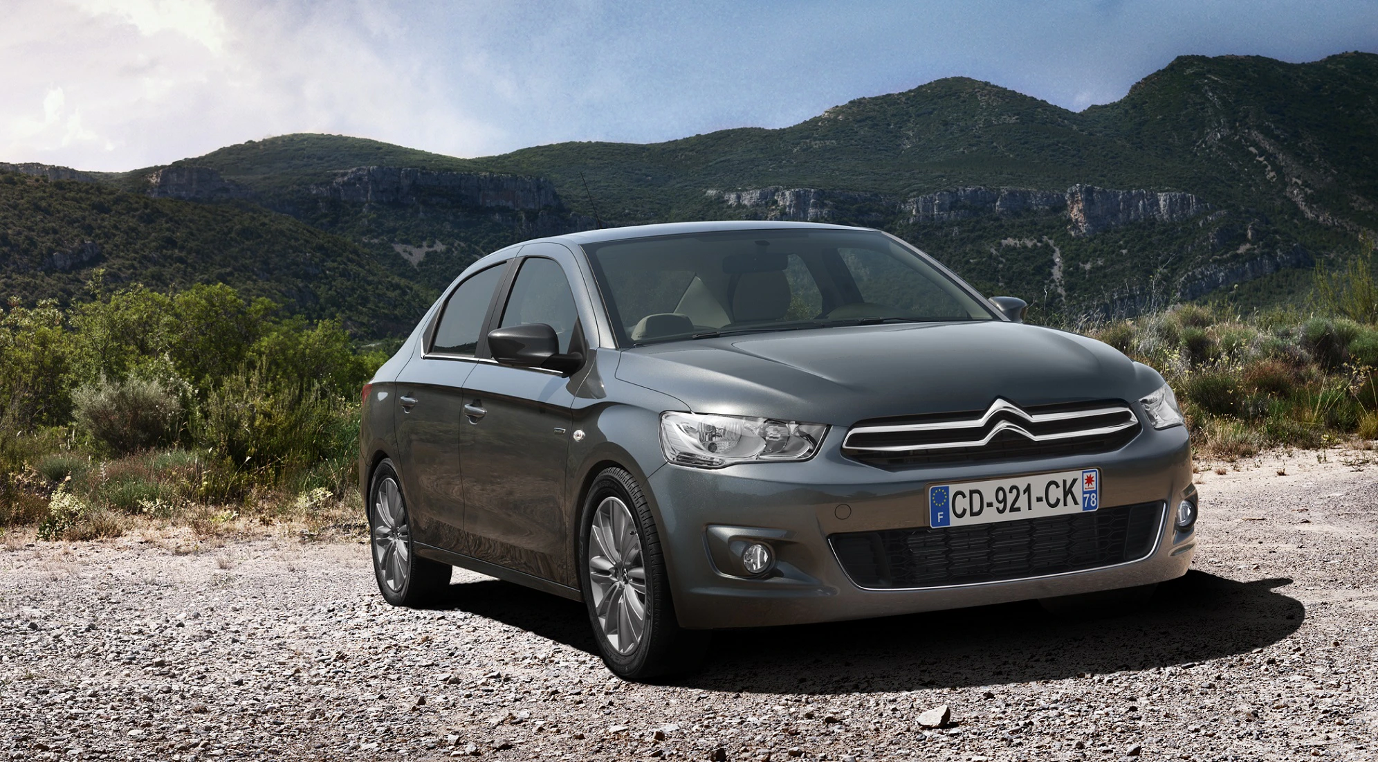 Silberner Citroën C-Elysée steht auf einem Schotterweg vor einer hügeligen Landschaft unter wolkigem Himmel.