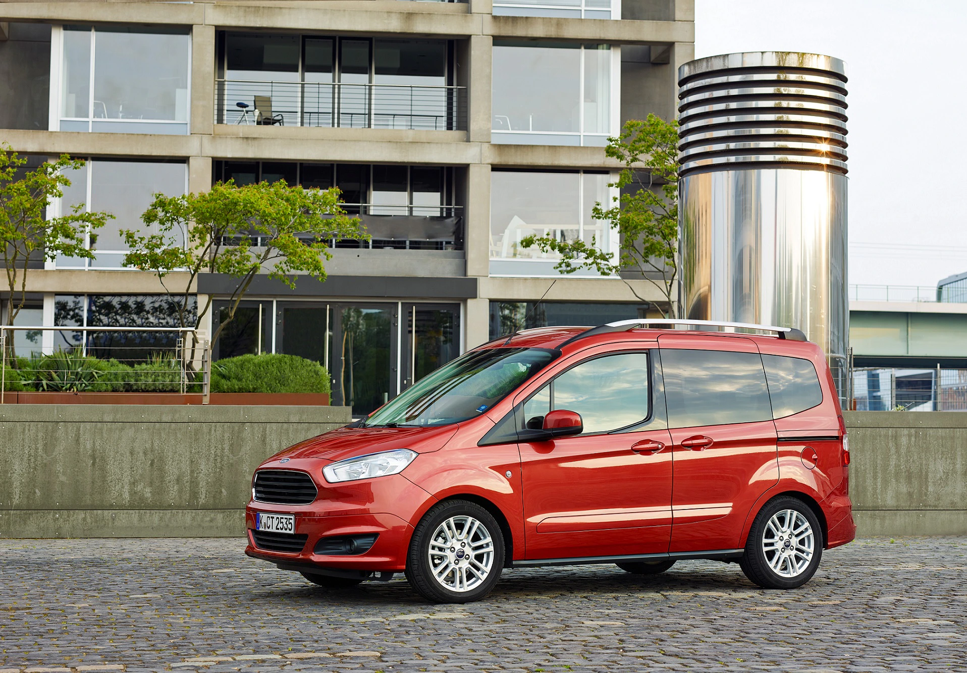 Roter Ford Tourneo Courier steht auf Kopfsteinpflaster vor einem modernen Gebäude und einem großen, silbernen Lüftungsschacht.