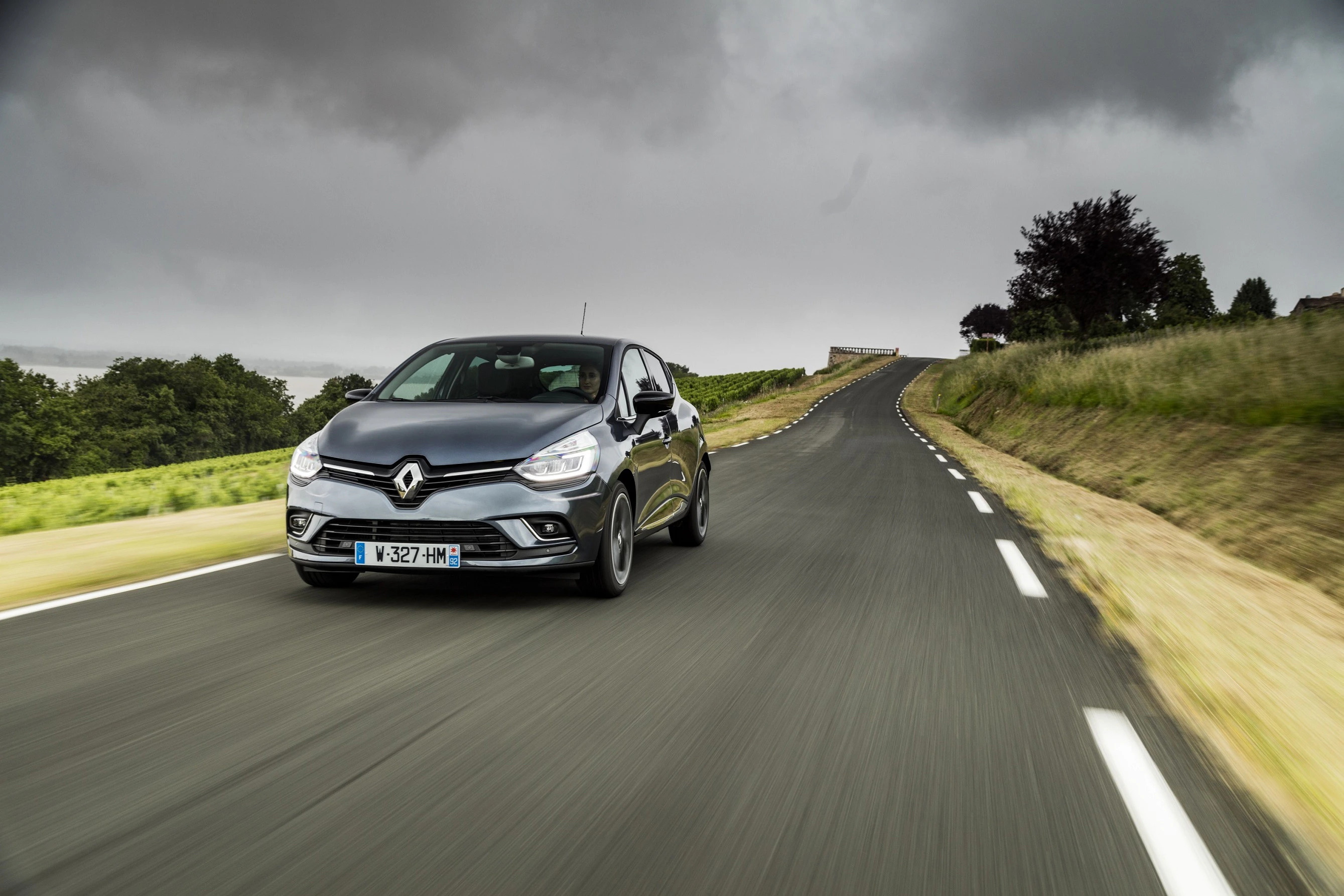Ein grauer Renault fährt auf einer kurvigen, zweispurigen Landstraße. Der Himmel ist bewölkt, und die Straße ist von Wiesen gesäumt.