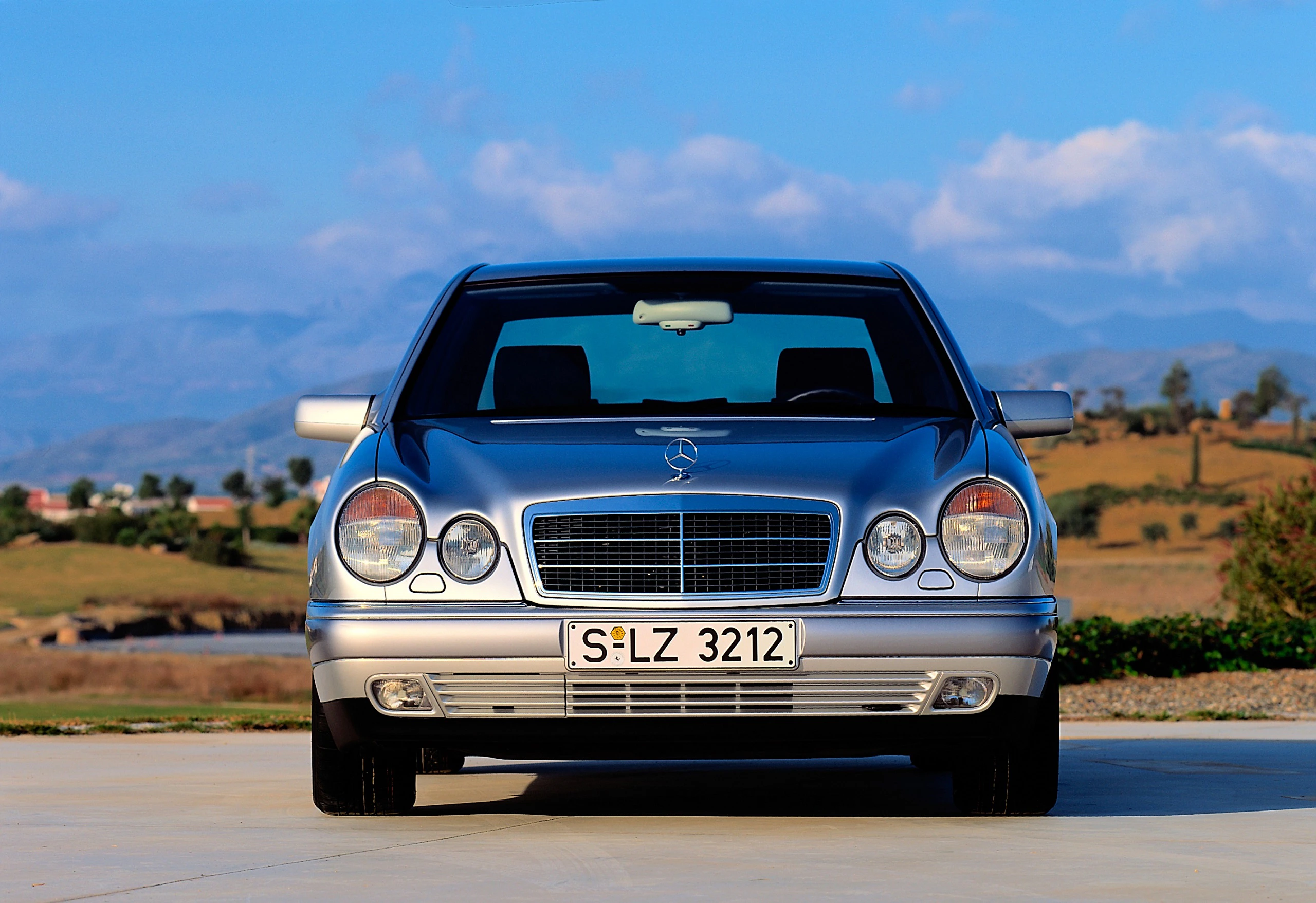Silberner Mercedes-Benz E-Klasse, geparkt auf einem sonnigen Platz mit Bergen und blauem Himmel im Hintergrund.