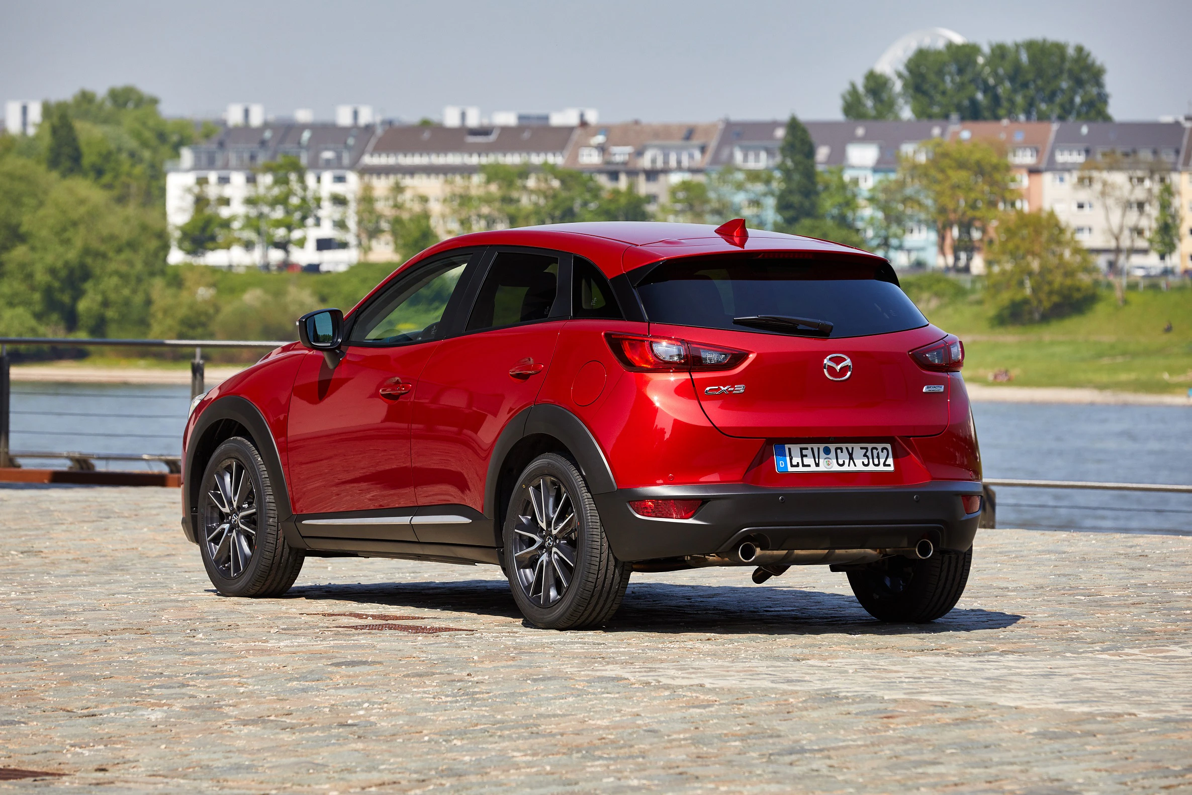 Roter Mazda CX-3 parkt auf gepflastertem Platz mit Fluss und Stadt im Hintergrund bei sonnigem Wetter.
