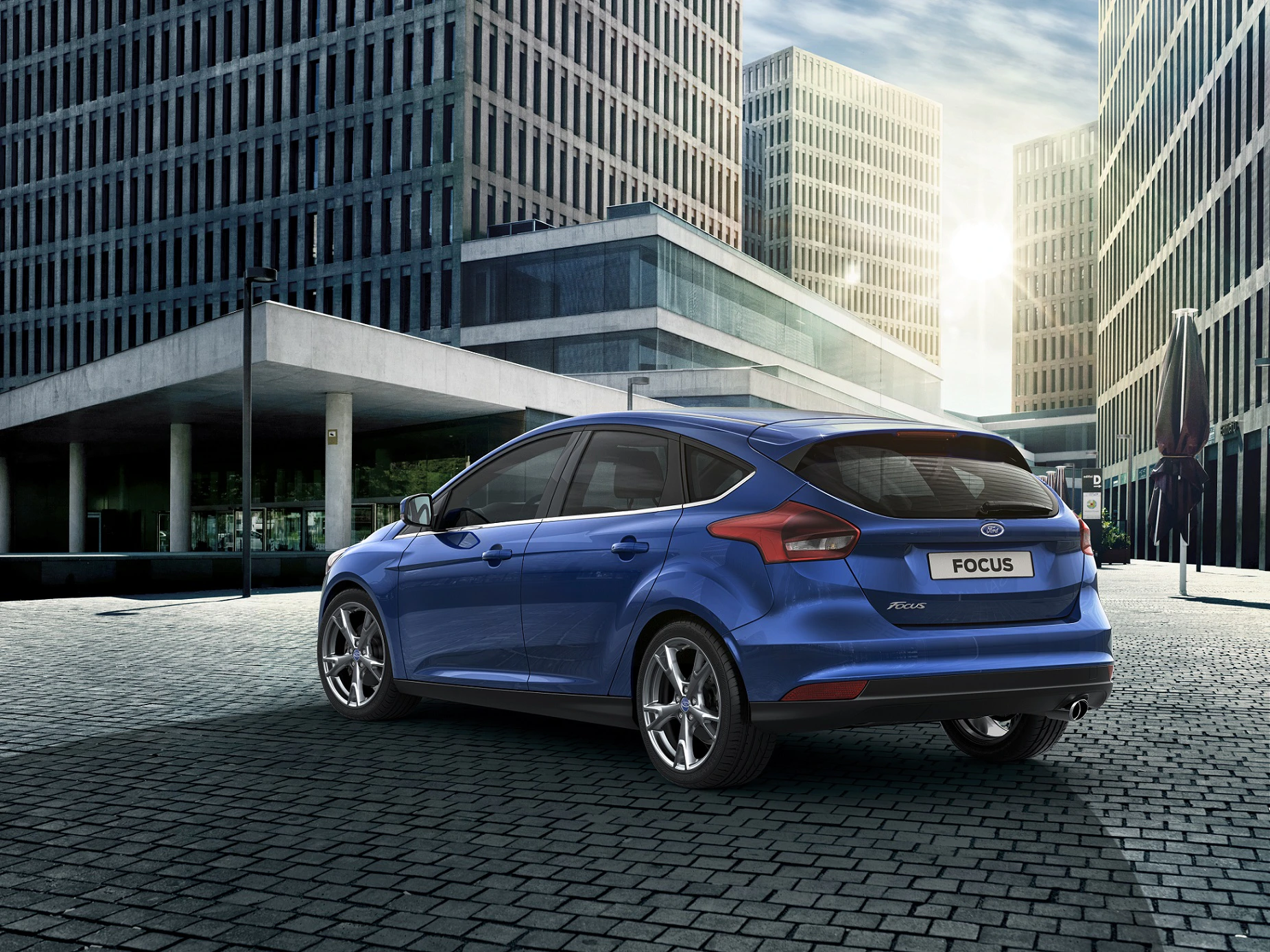 Blauer Ford Focus parkt auf gepflastertem Platz, umgeben von modernen Bürogebäuden und unter bewölktem Himmel.