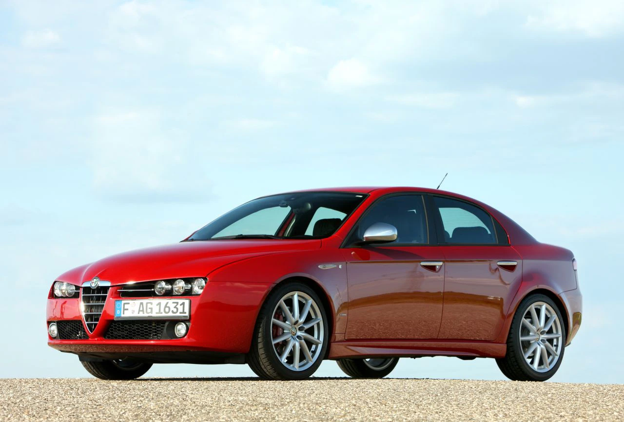 Roter Alfa Romeo 159, seitlich fotografiert, geparkt auf einem steinigen Untergrund vor bewölktem Himmel.