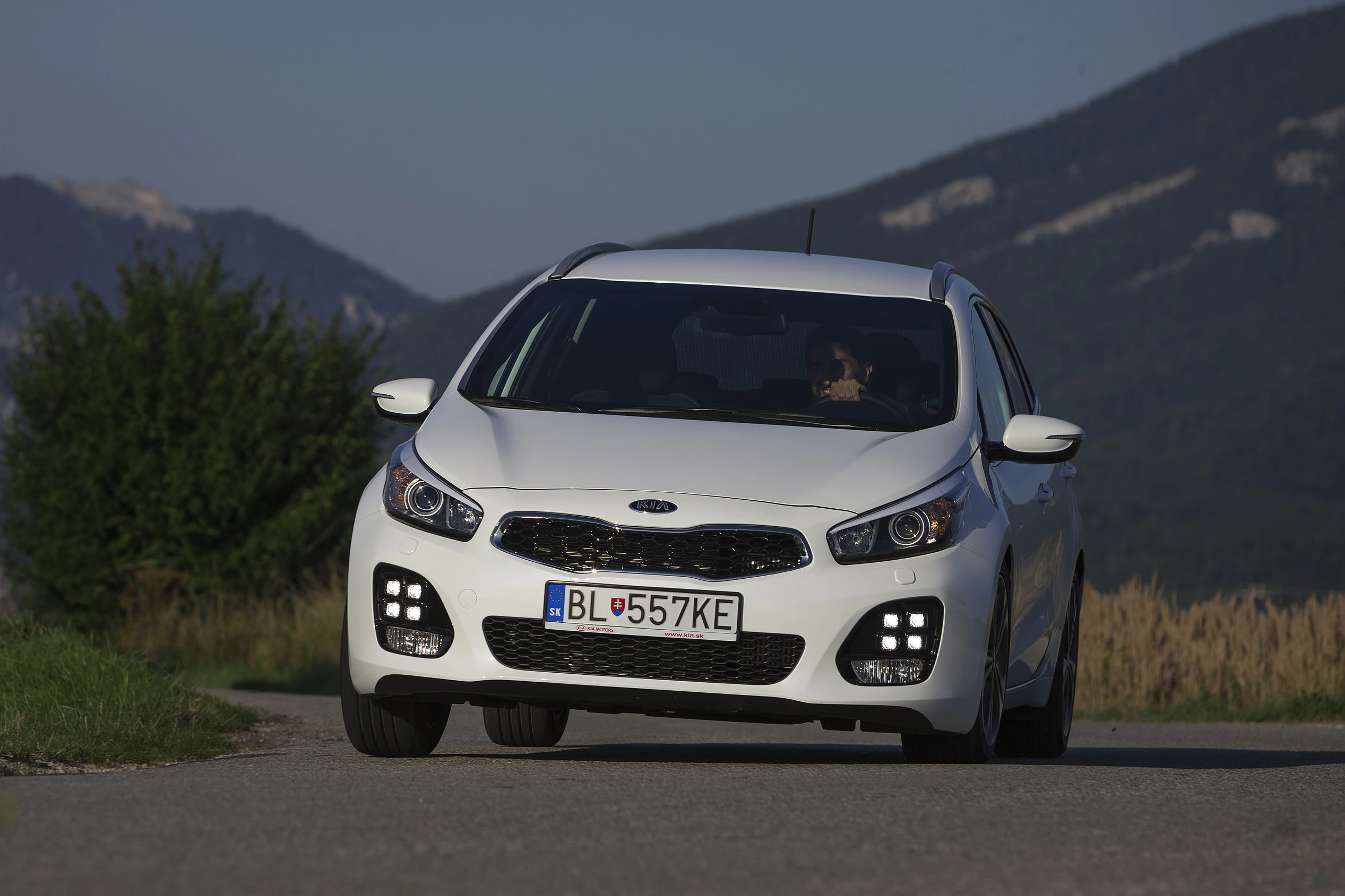 Weißes Auto fährt auf einer Landstraße vor bergiger Kulisse. Fahrzeug mit LED-Lichtern und Nummernschild BL 557KE.