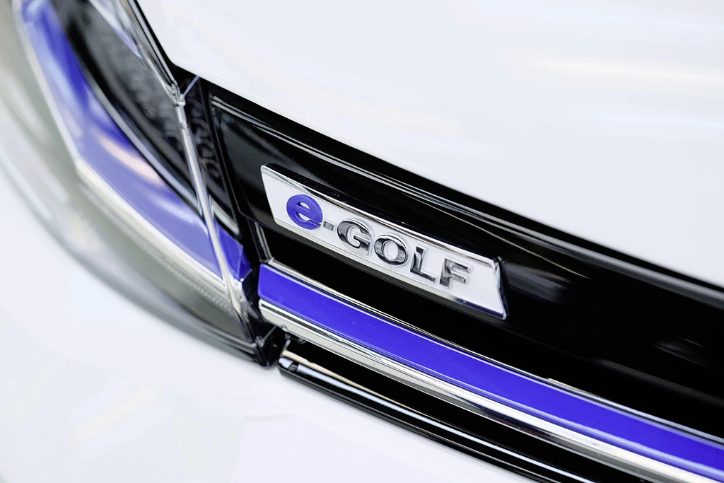Nahaufnahme des Emblems eines VW e-Golf auf einem weißen Auto, mit blauer Linie am Kühlergrill.