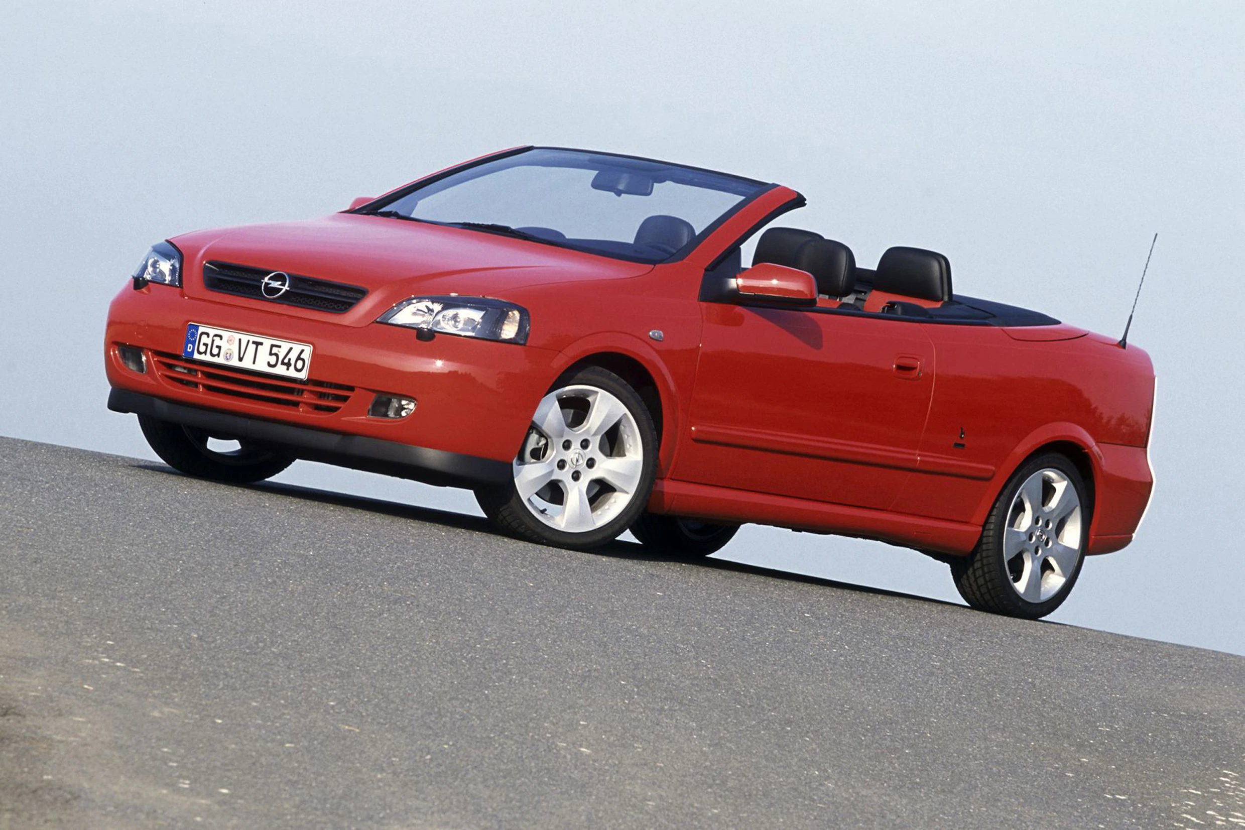 Rotes Opel Cabrio fährt auf einer Straße, klarer Himmel im Hintergrund. Fahrzeug steht leicht schräg, Dach geöffnet.