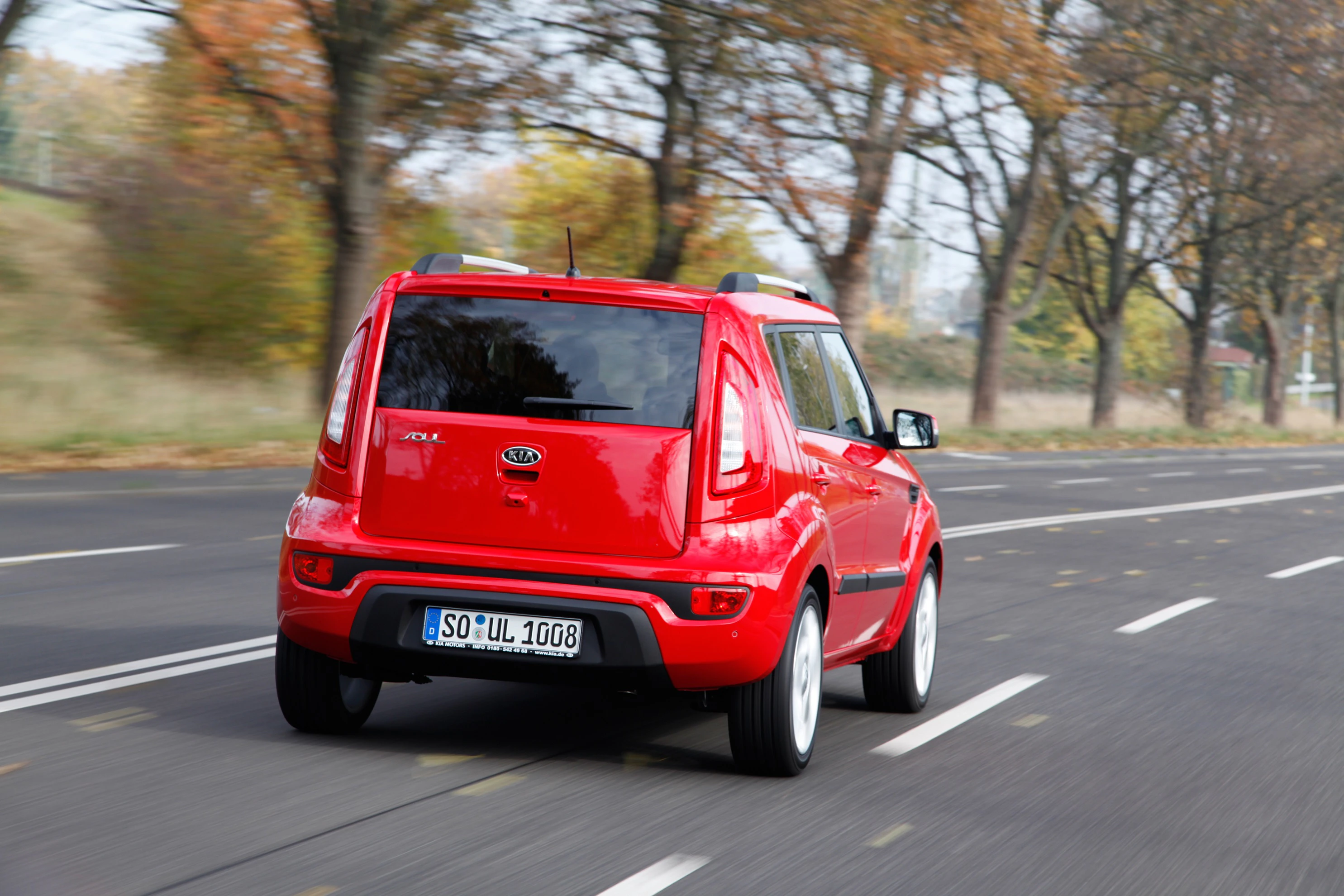 Roter Kia Soul fährt auf einer Landstraße im Herbst. Bäume mit buntem Laub sind im Hintergrund zu sehen.