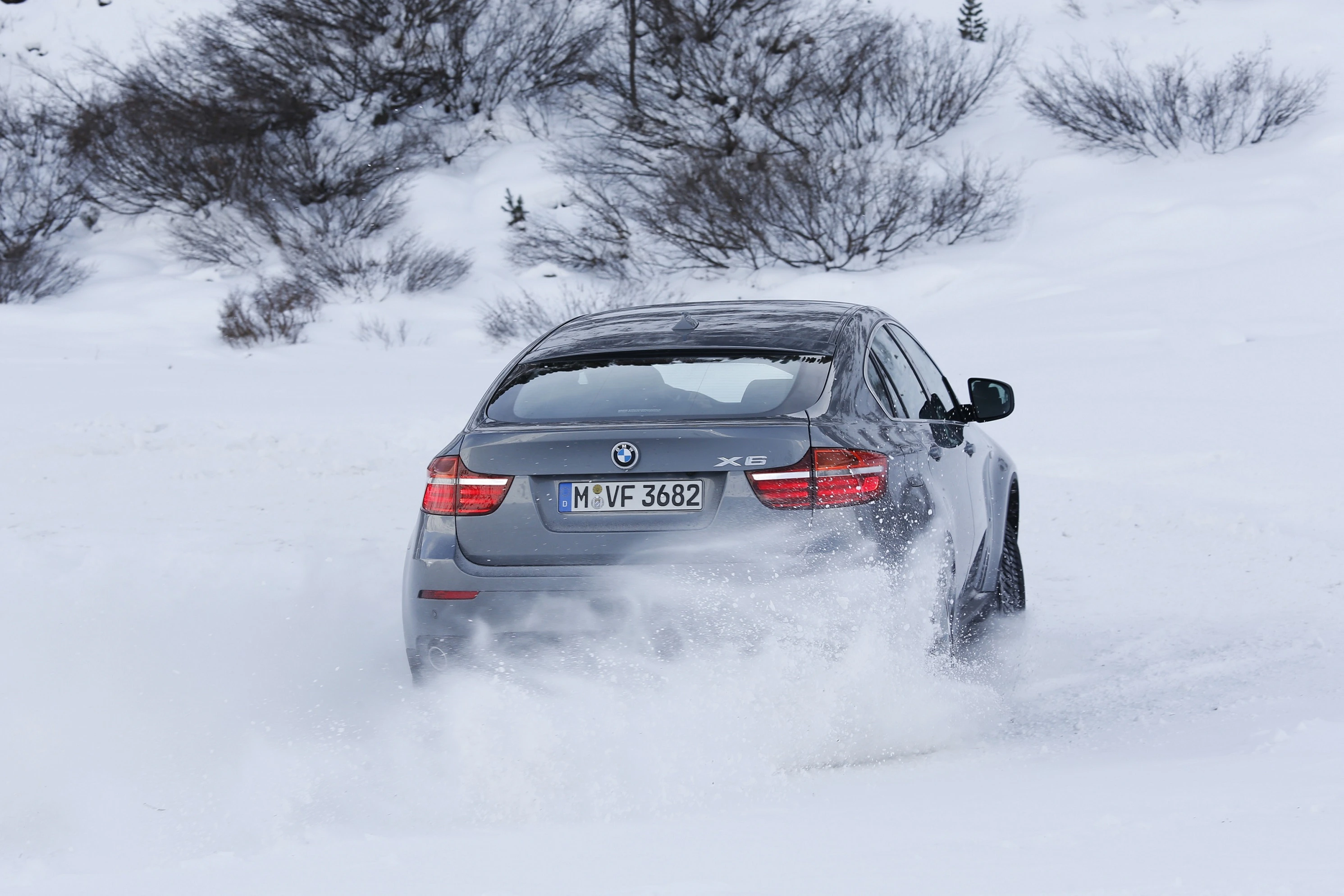 Ein grauer BMW X6 fährt durch verschneite Landschaft und hinterlässt eine Spur im Schnee, während Bäume den Hintergrund säumen.