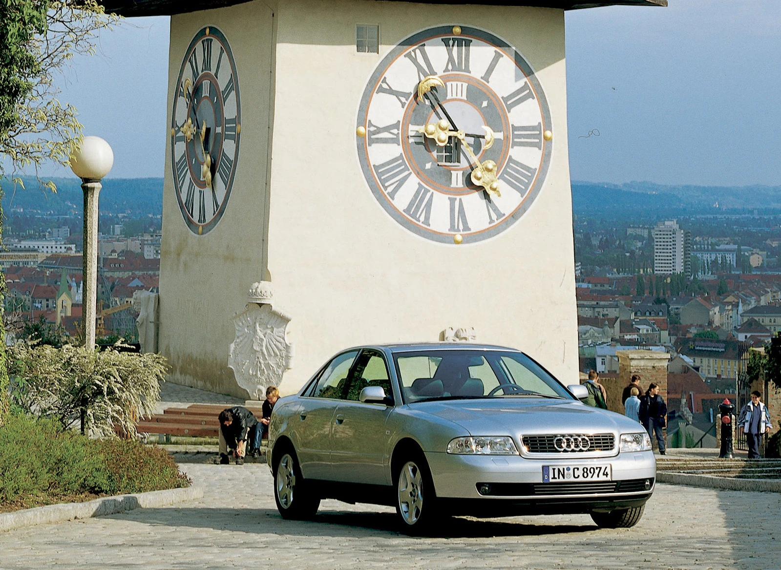 Silbernes Auto steht vor dem Grazer Uhrturm, einem berühmten Wahrzeichen mit großen Uhren und Stadtpanorama im Hintergrund.