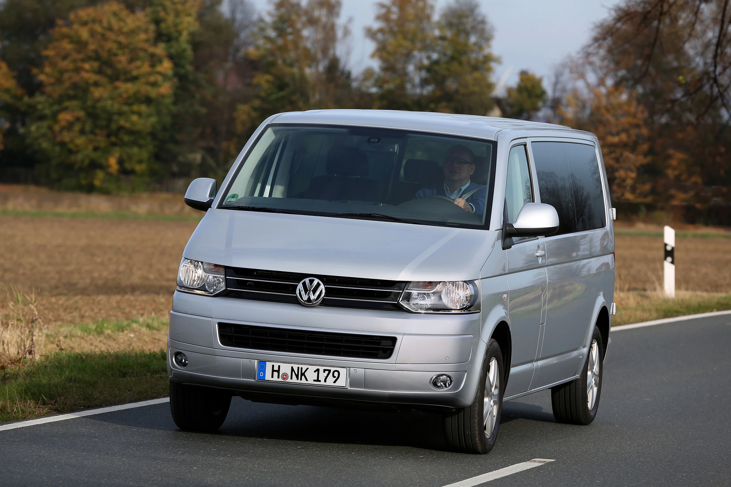 Silberner VW Transporter fährt auf einer Landstraße, herbstliche Bäume im Hintergrund.