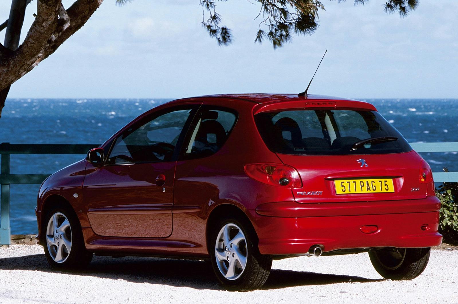 Roter Peugeot 206 an Küstenstraße geparkt, Meer im Hintergrund, sonniges Wetter, Baumzweige über dem Auto.