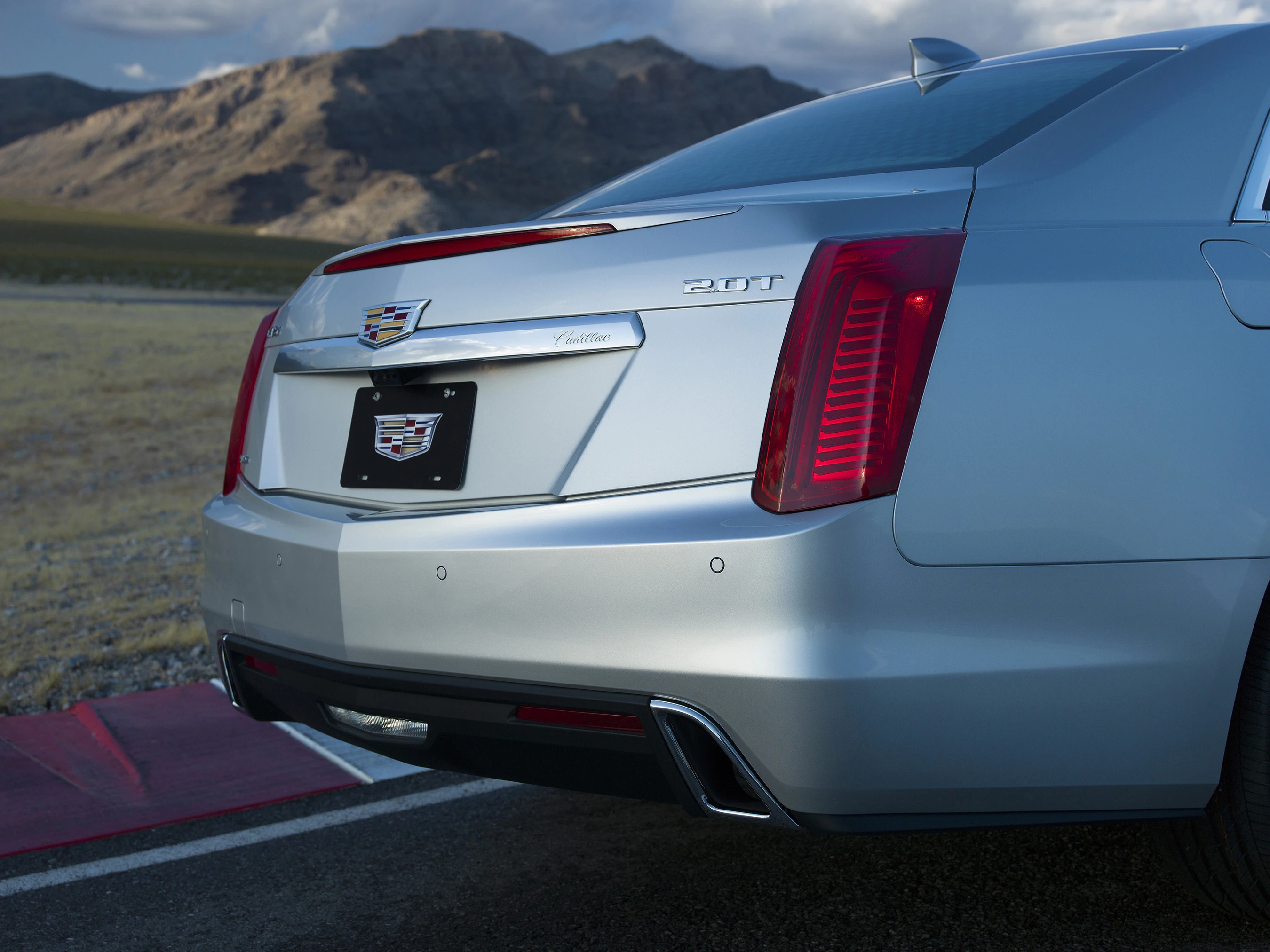 Rückansicht eines silbernen Cadillac 2.0T auf einer Straße mit Bergen im Hintergrund. Kein Kennzeichen am Fahrzeug.