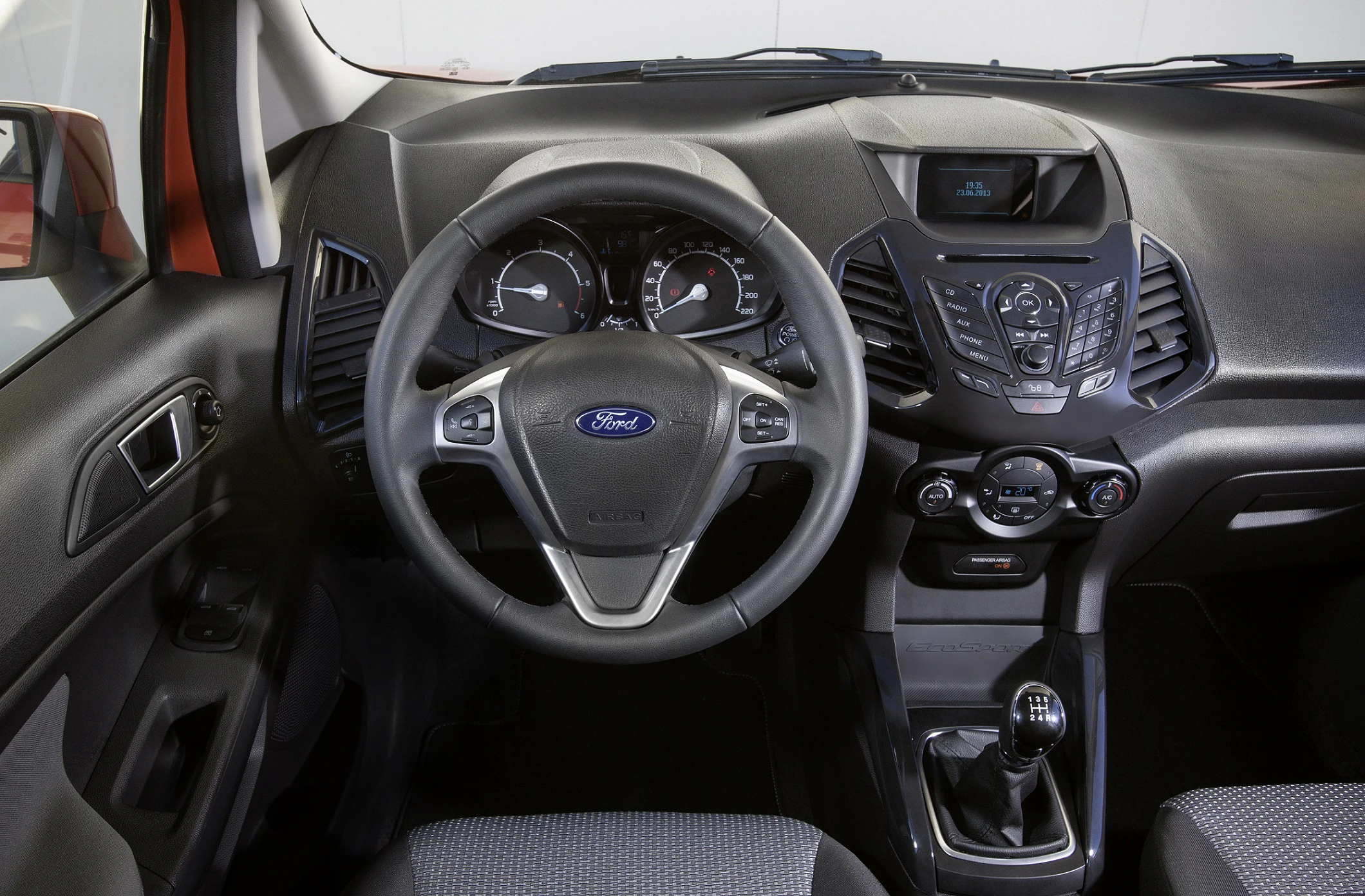 Innenraum eines Autos mit Ford-Logo auf dem Lenkrad, Armaturenbrett mit Tasten und Drehknöpfen, analogen Anzeigen und manueller Schaltknüppel.
