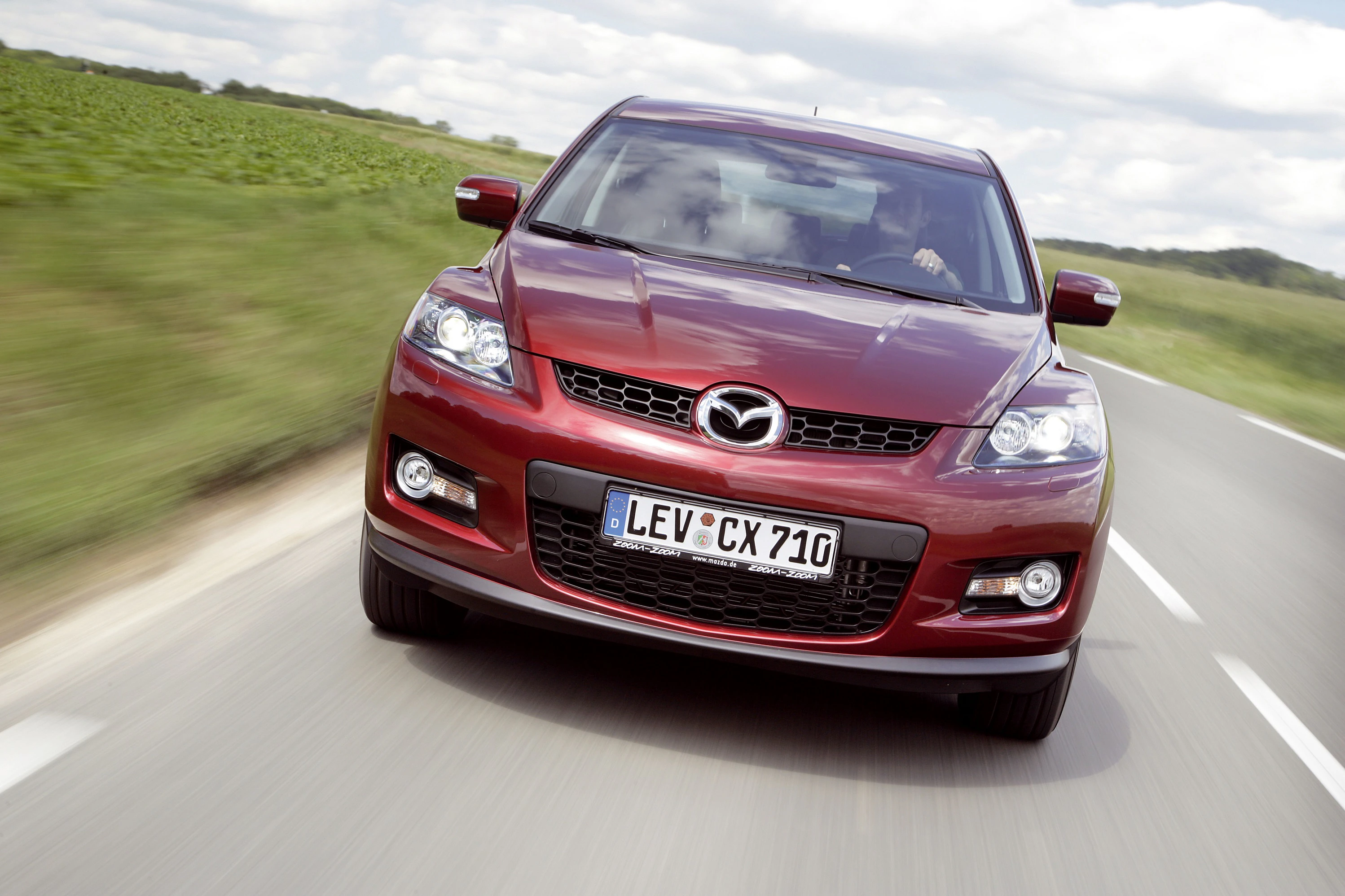 Roter Mazda CX-7 fährt auf Landstraße, mit grünem Feld und blauem Himmel im Hintergrund.