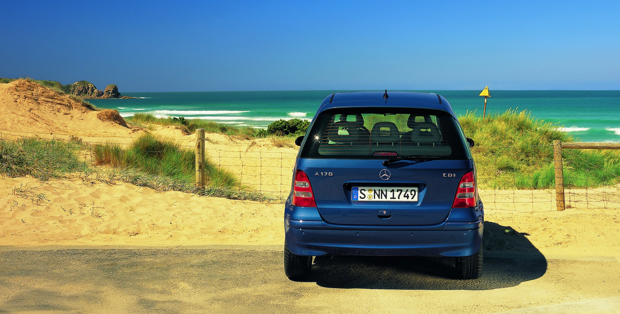 Blauer Mercedes A170 steht auf sandiger Straße nahe einer Düne mit Blick auf das Meer und einen klaren blauen Himmel.
