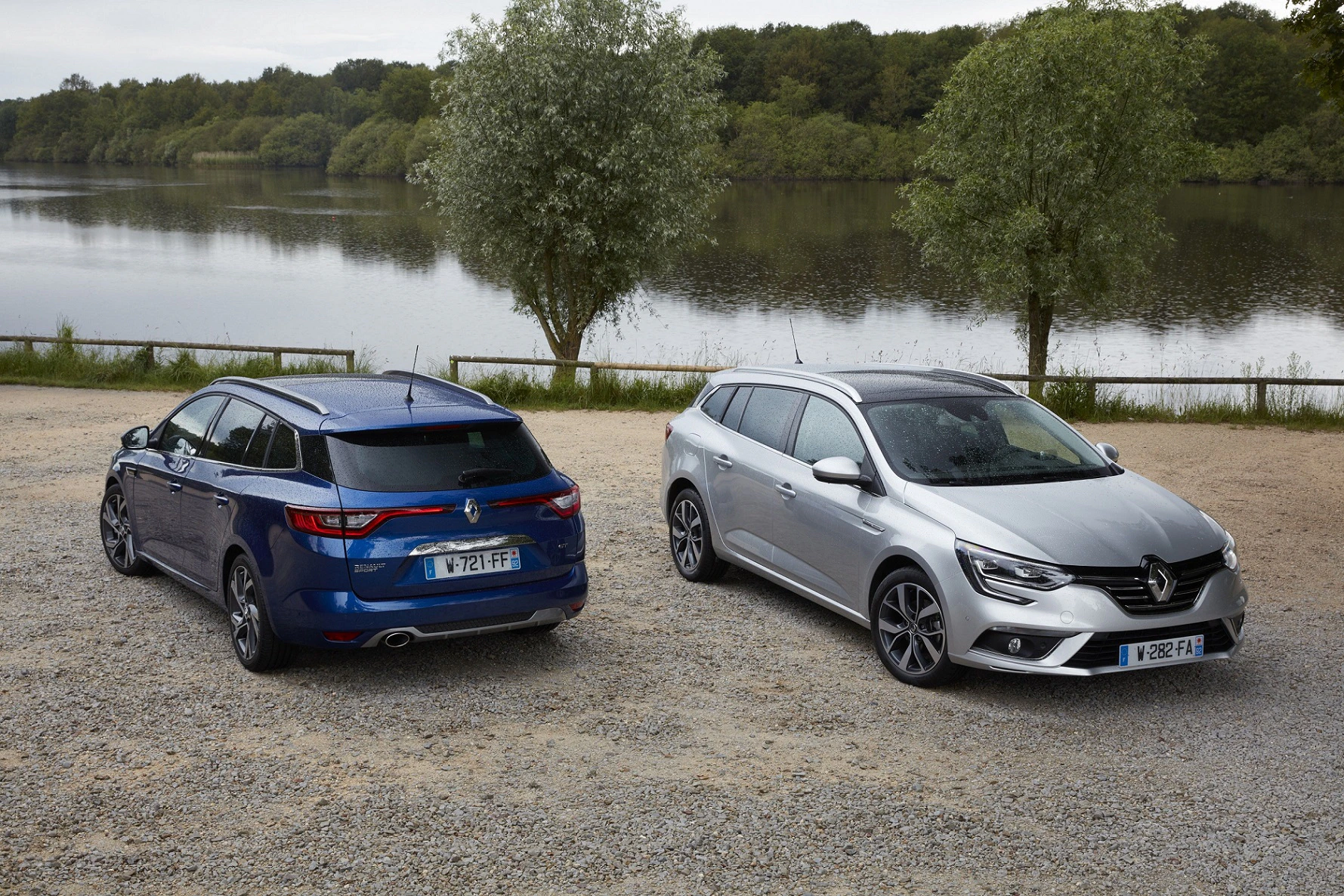 Zwei geparkte Autos, ein blaues und ein silbernes, an einem Kiesweg vor einem See mit Bäumen im Hintergrund.