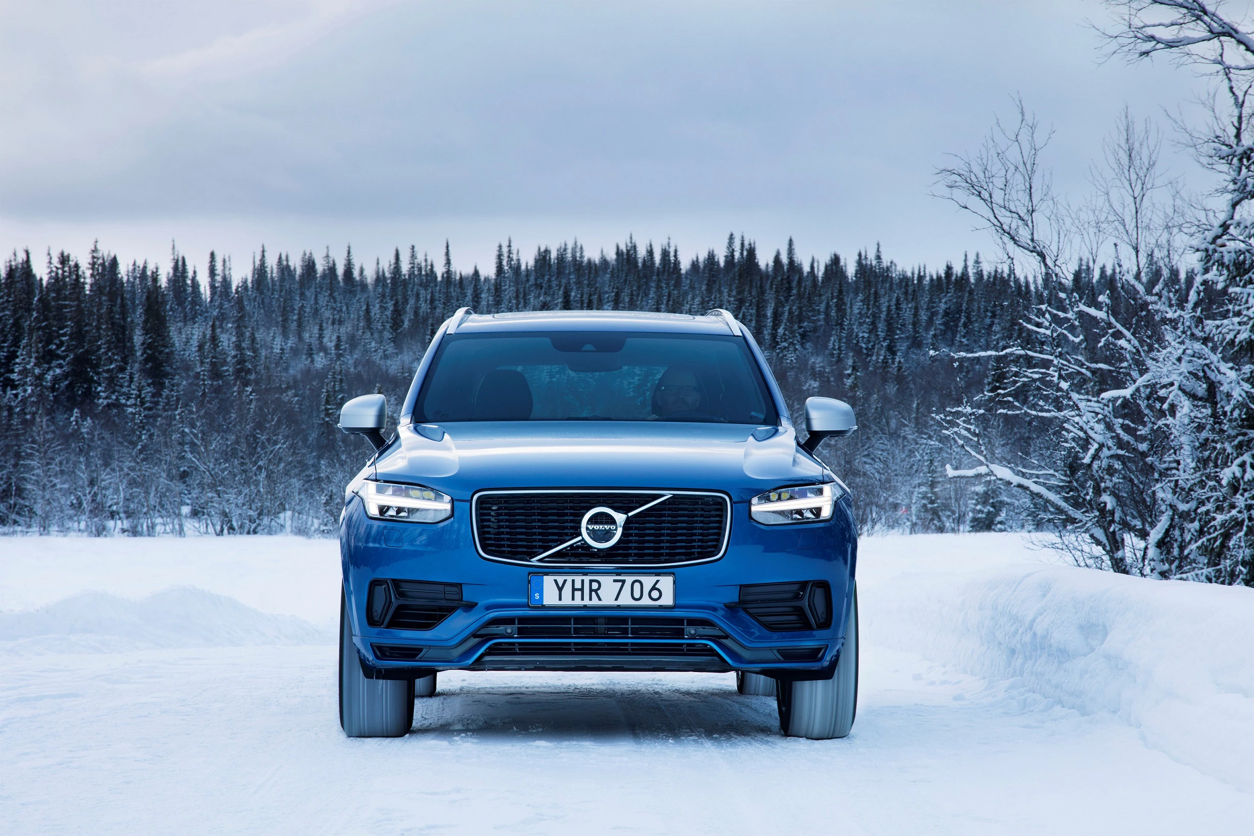 Blauer Volvo SUV fährt auf verschneiter Straße, umgeben von schneebedeckten Bäumen vor einem bewölkten Himmel.