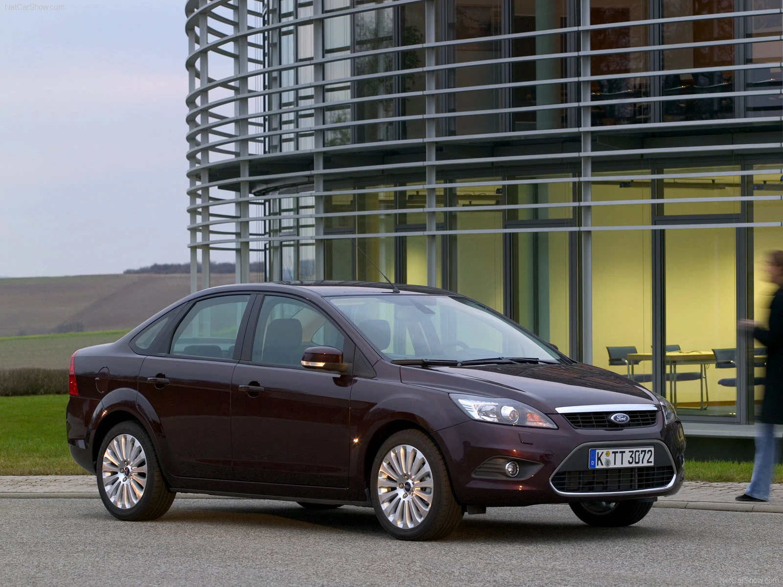 Dunkelvioletter Ford Focus Limousine vor einem modernen Glasgebäude, mit einer unscharfen Person im Hintergrund.