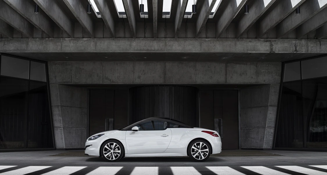Weißes Sportcoupé vor moderner, betonlastiger Architektur mit markanten Linien und symmetrischen Öffnungen im Hintergrund.