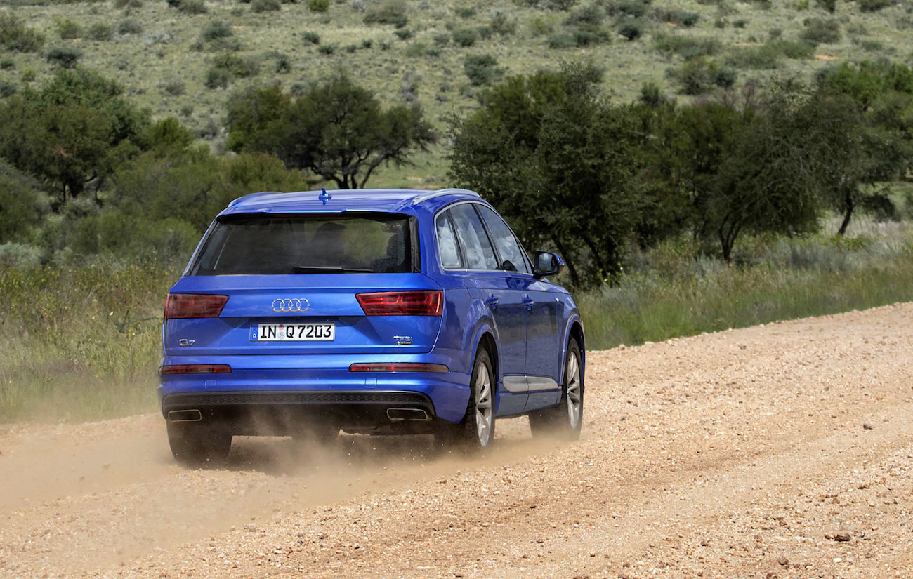 Blauer Audi SUV fährt auf einer staubigen Landstraße. Im Hintergrund sind Bäume und eine grasbewachsene Landschaft zu sehen.