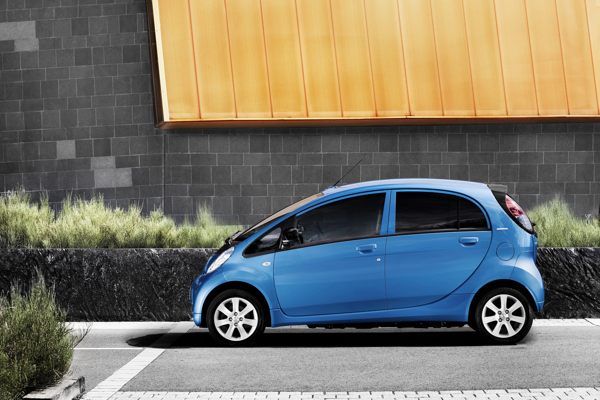 Kompaktes blaues Auto parkt vor einer modernen Mauer mit grauen und gelben Elementen, umgeben von grünen Pflanzen.
