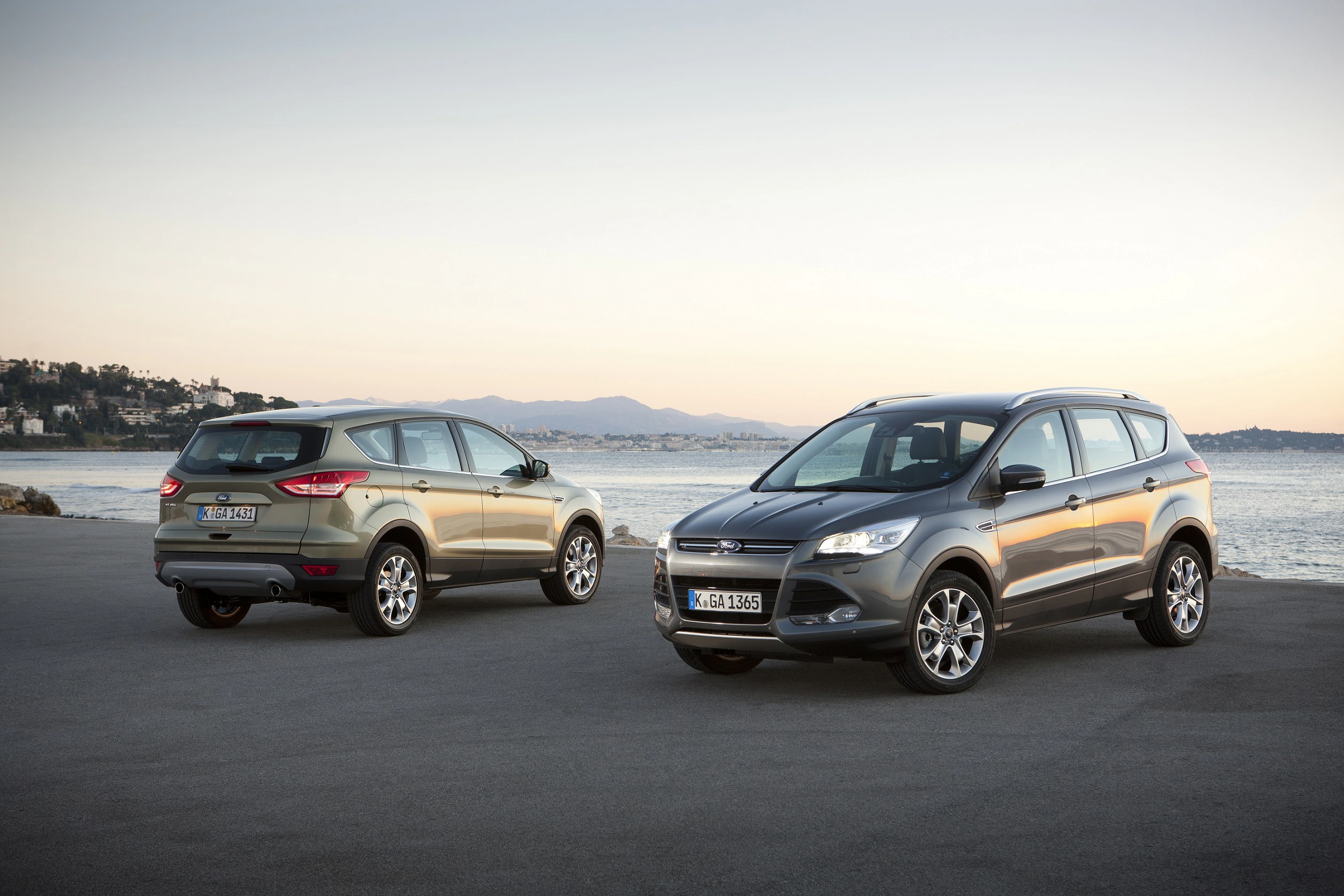 Zwei SUVs parken am Meer bei Sonnenuntergang. Der Himmel ist klar, und im Hintergrund sind Hügel und Gebäude zu sehen.