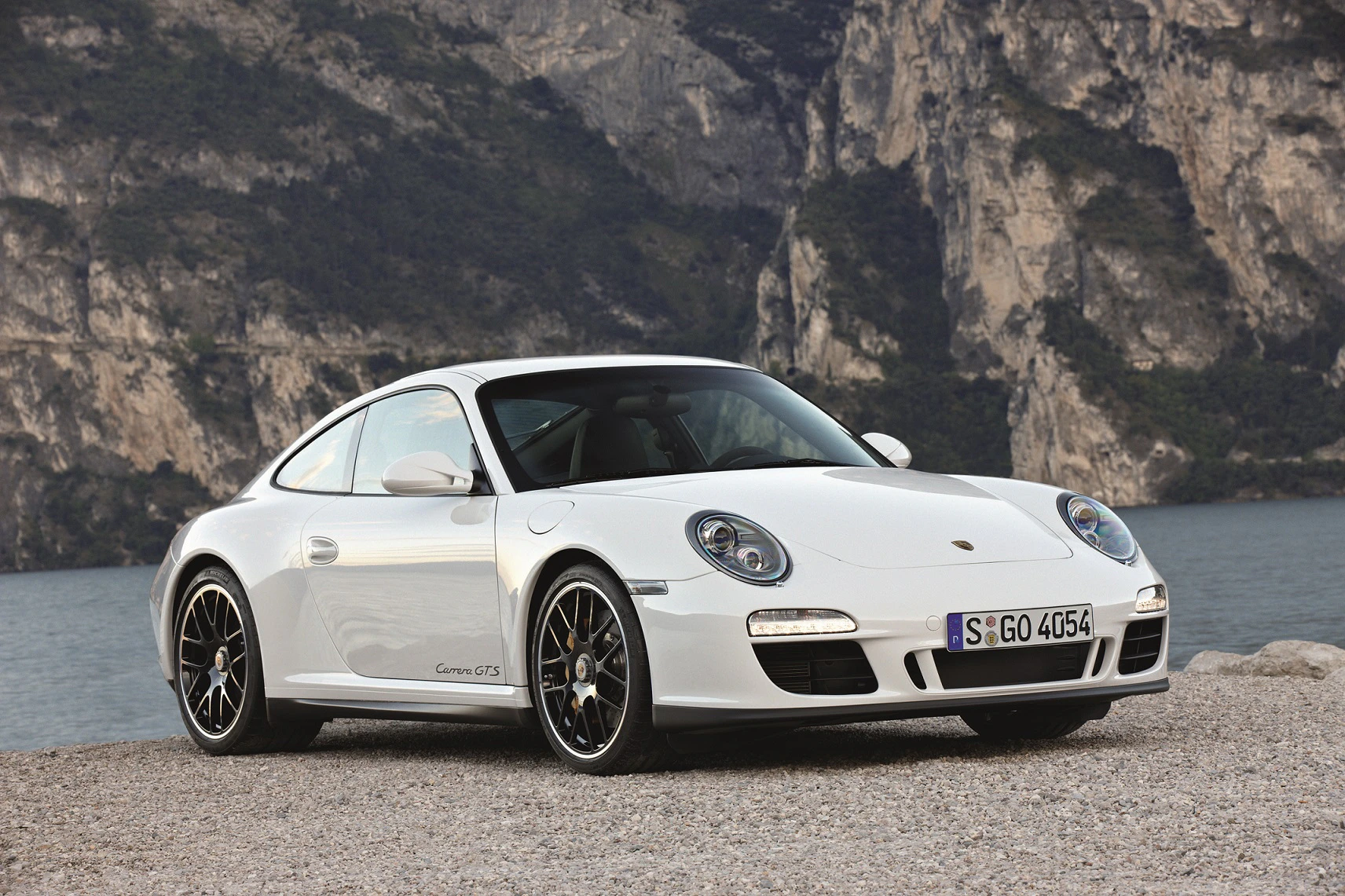 Weißer Sportwagen Porsche 911 Carrera GTS steht vor einer natürlichen Felswand und einem See im Hintergrund.