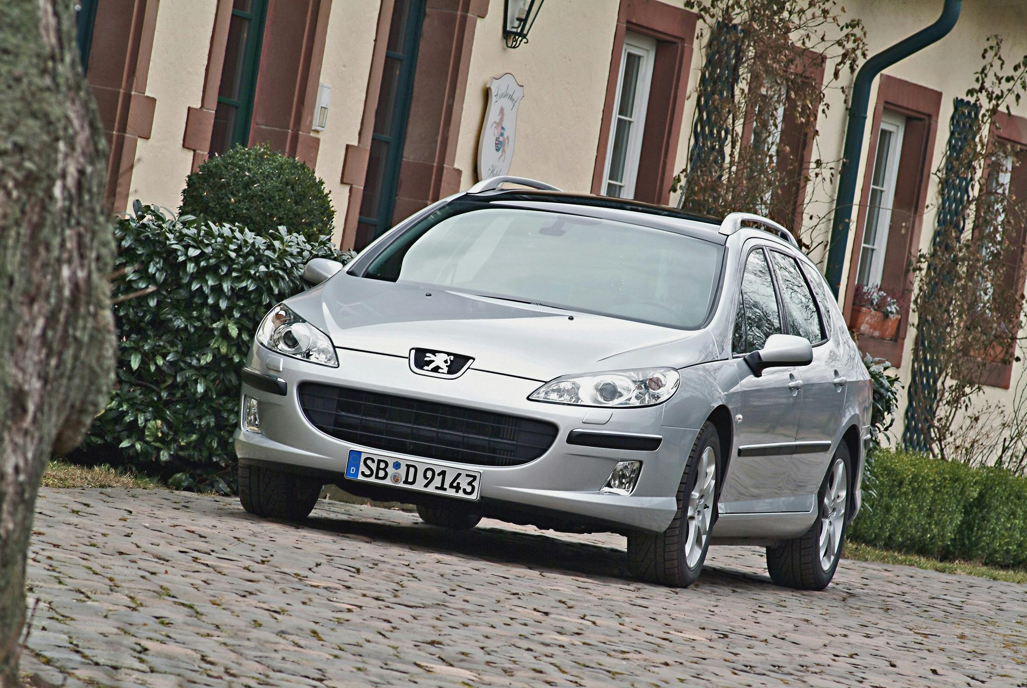 Silberner Peugeot-Kombi parkt auf einem gepflasterten Hof vor einem Gebäude mit roten Fensterrahmen und grüner Hecke.