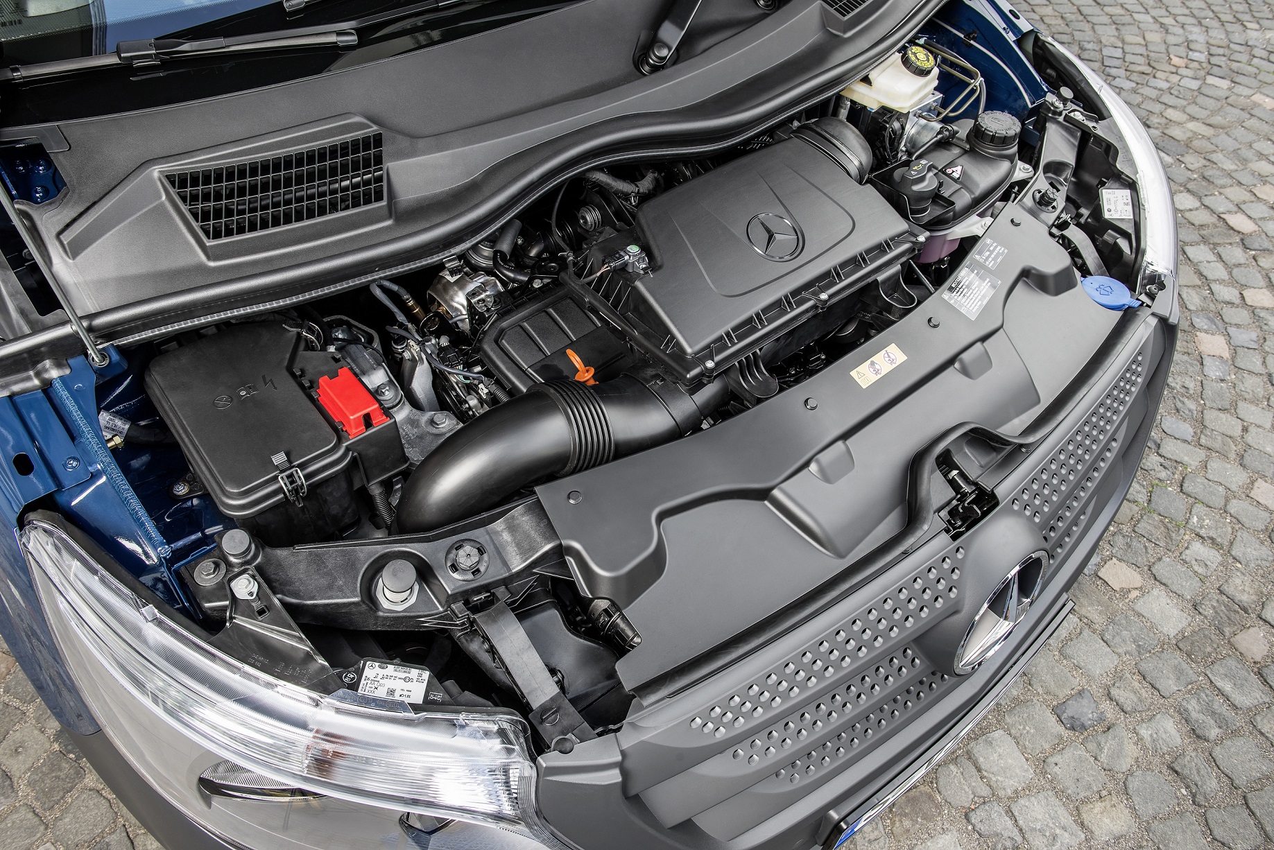 Motorraum eines Mercedes-Fahrzeugs, gezeigt von oben. Sichtbar sind der Motorblock, verschiedene Kabel und Rohrleitungen. Fahrzeug steht auf Kopfsteinpflaster.