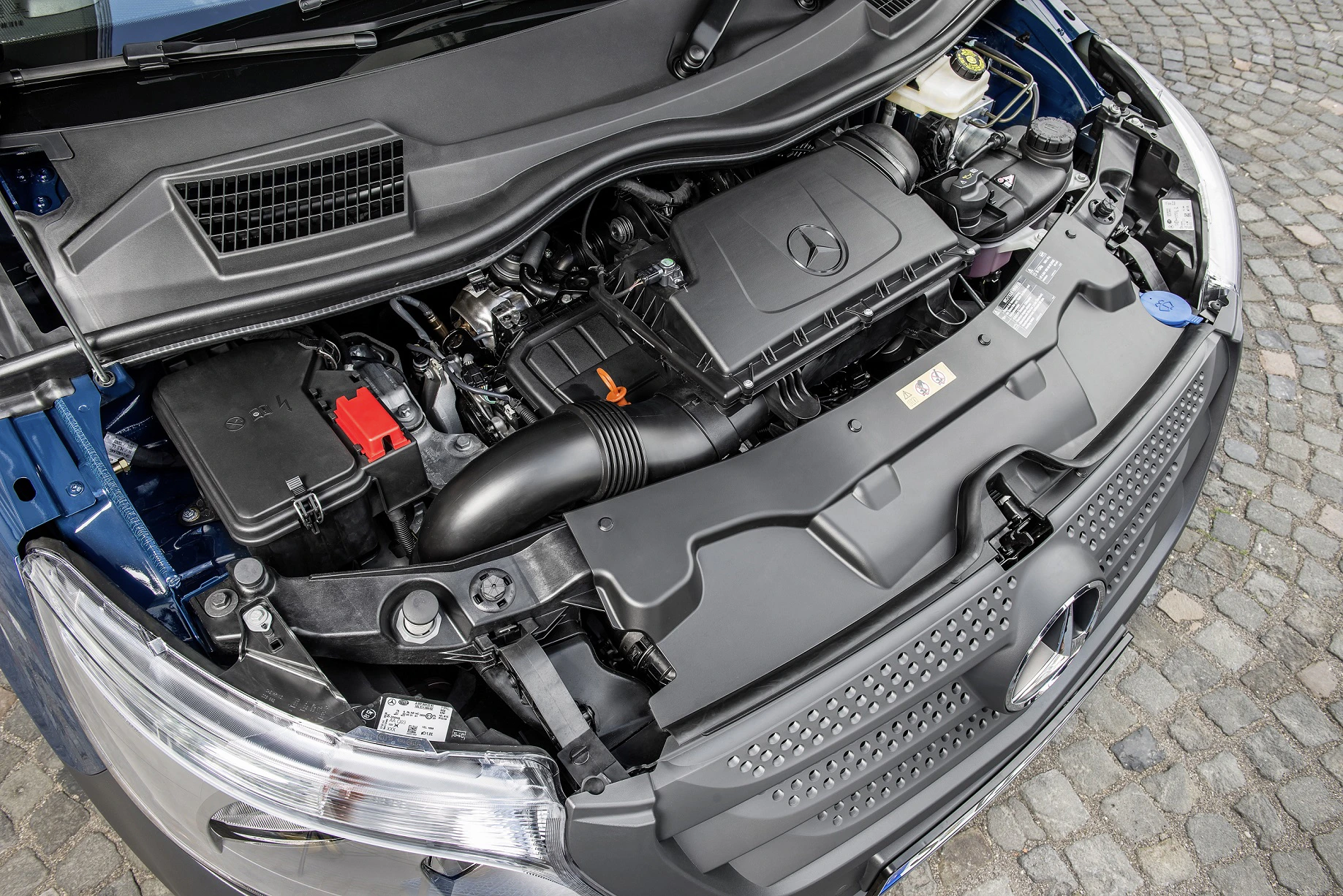 Motorraum eines Mercedes-Fahrzeugs, gezeigt von oben. Sichtbar sind der Motorblock, verschiedene Kabel und Rohrleitungen. Fahrzeug steht auf Kopfsteinpflaster.