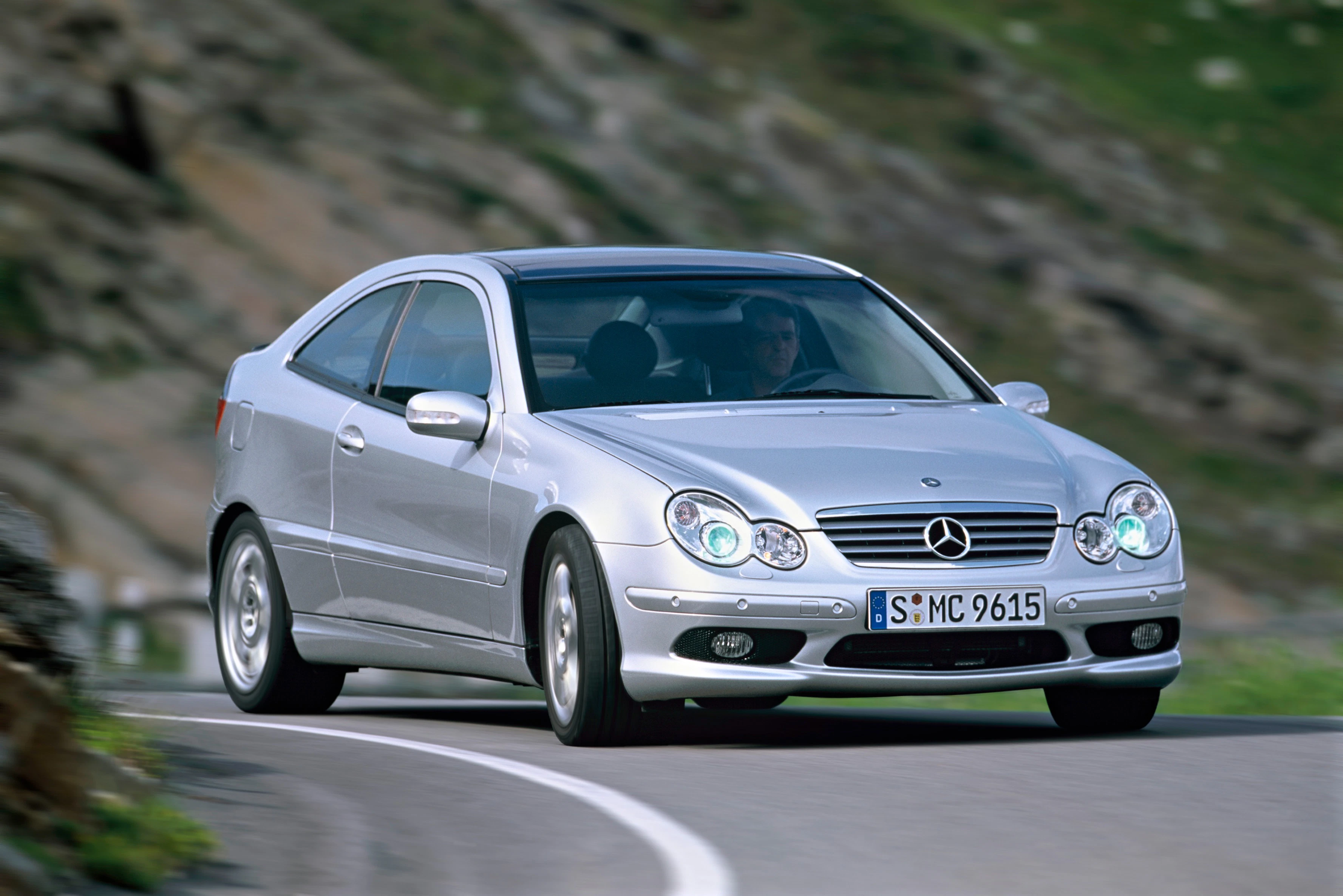 Silberner Mercedes-Benz CLK fährt auf kurviger Bergstraße. Fahrer im Auto, grüne Landschaft im Hintergrund.