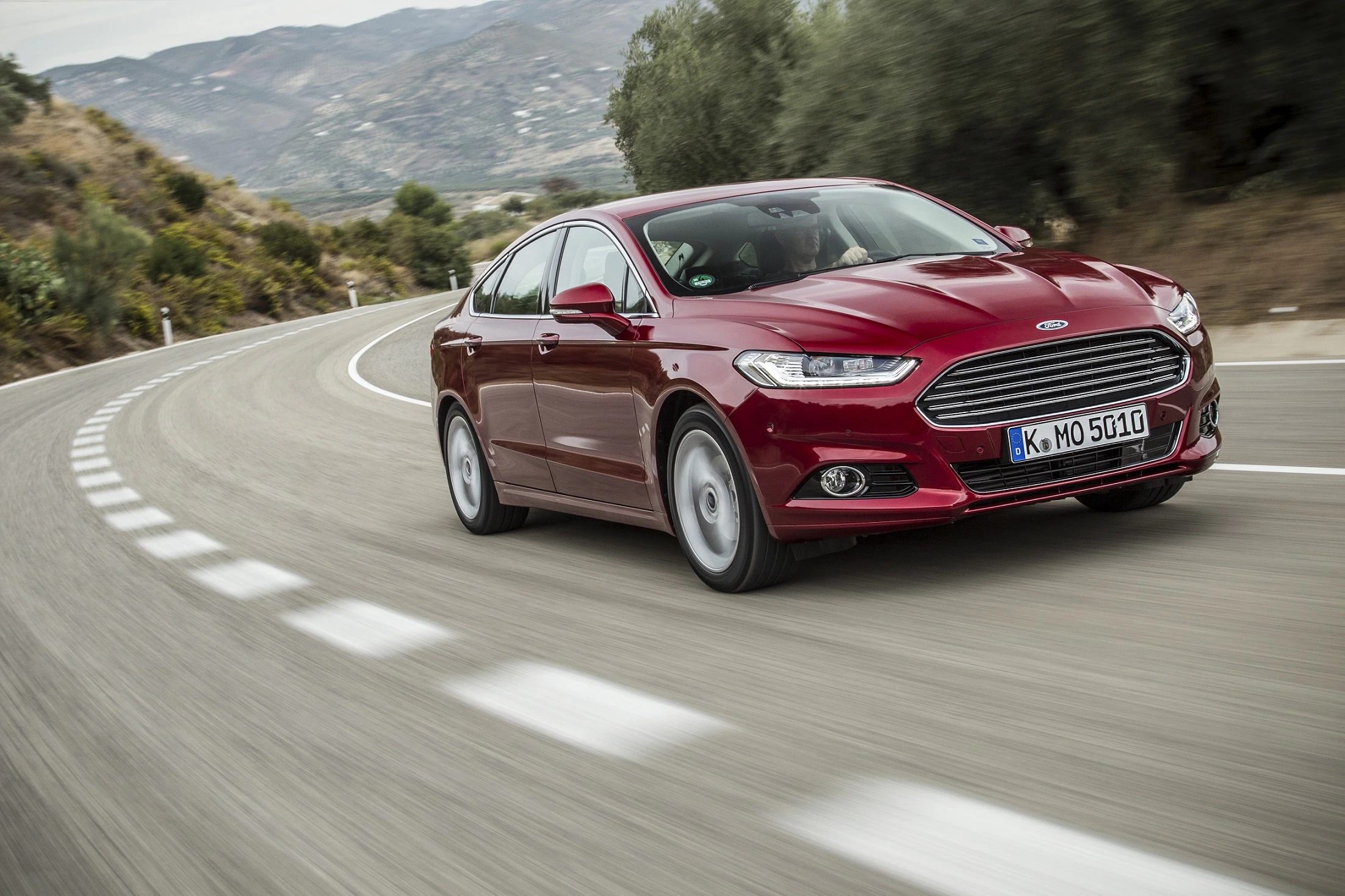 Roter Ford auf einer kurvigen, ländlichen Straße mit Bäumen und Bergen im Hintergrund unterwegs.