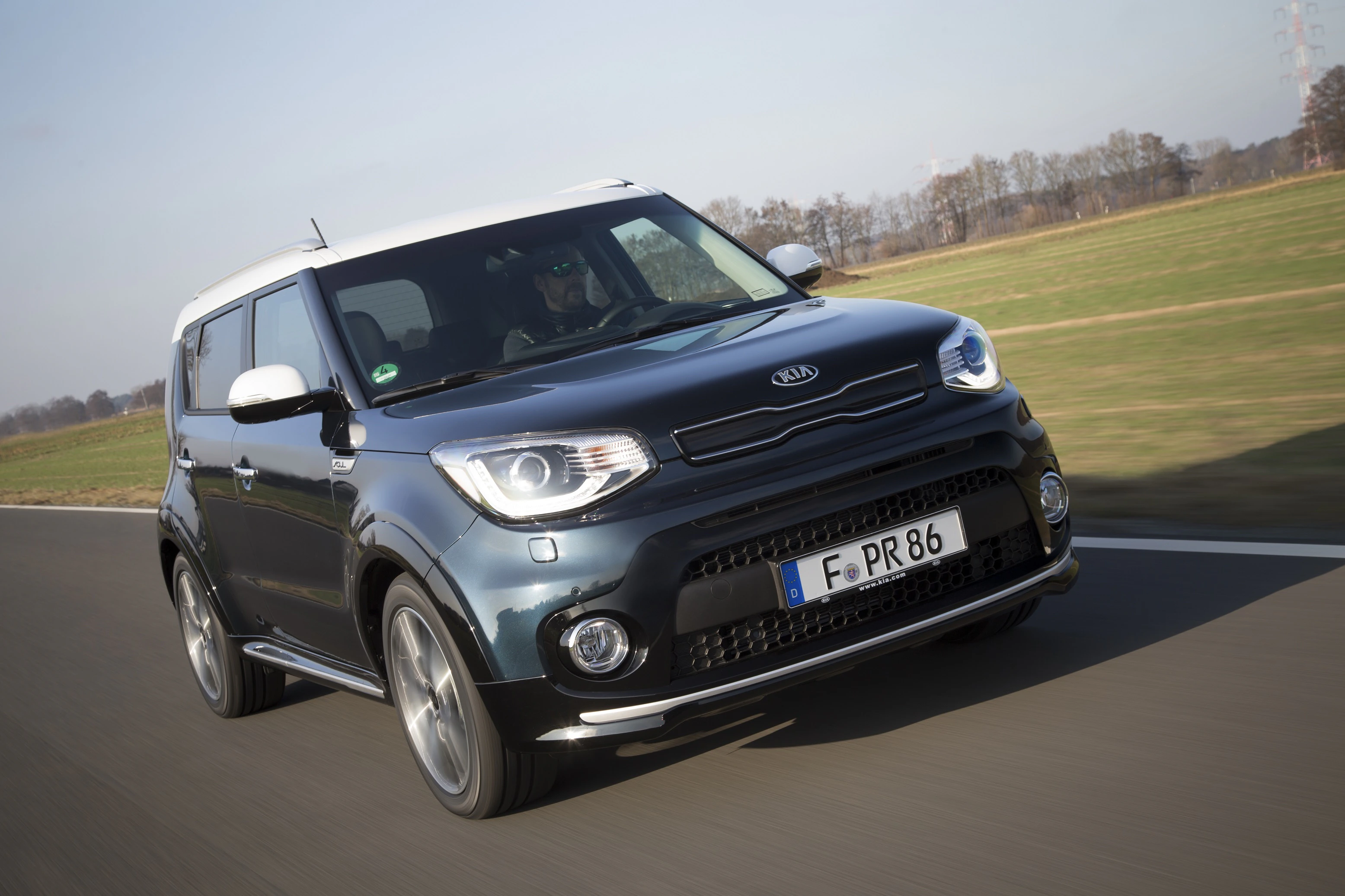 Ein schwarzer Kia Soul fährt auf einer Landstraße. Im Hintergrund sind Bäume und ein klarer Himmel zu sehen.