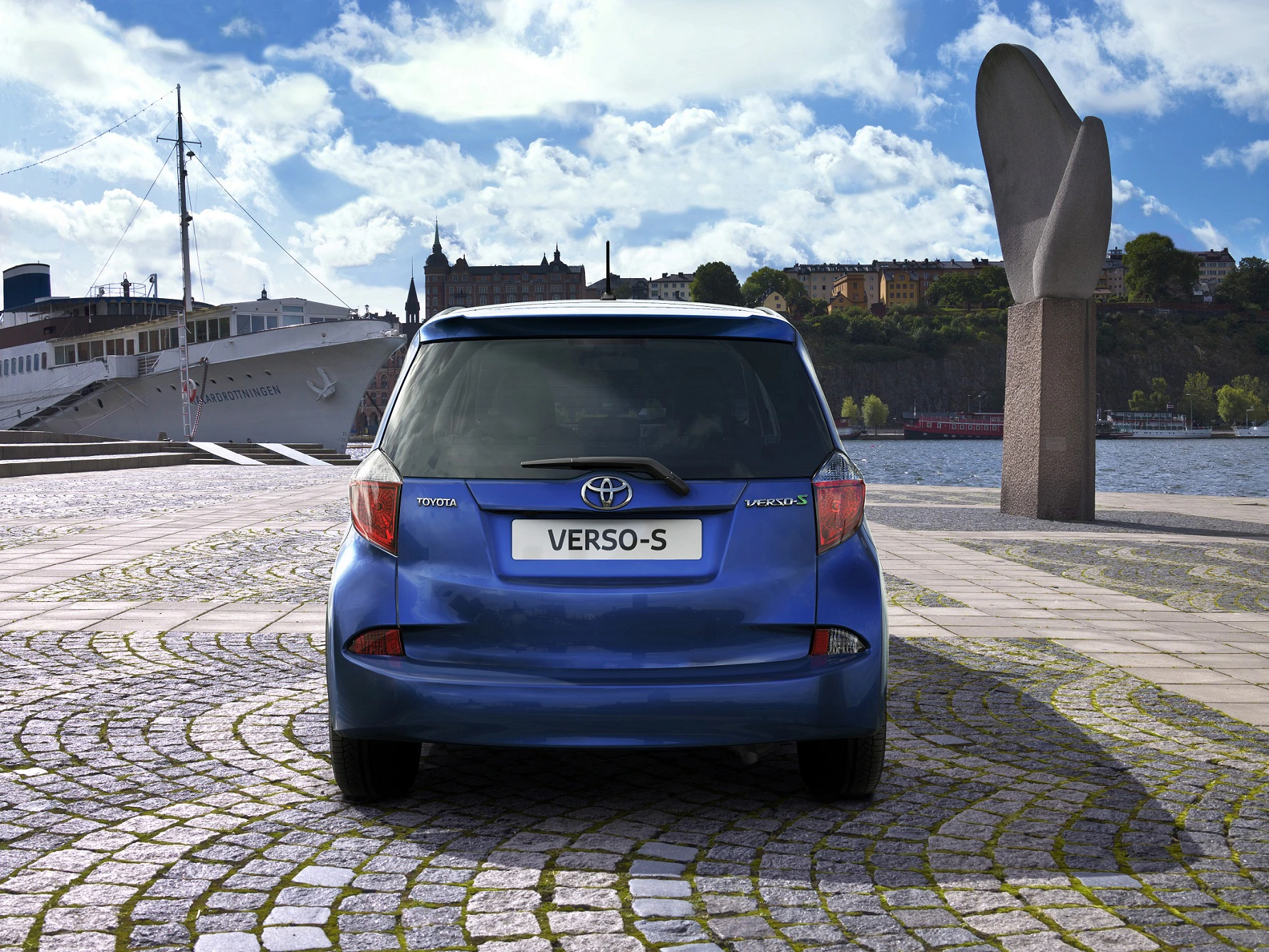 Blauer Toyota Verso-S geparkt auf gepflastertem Platz am Wasser, im Hintergrund ein Schiff und Stadtansicht unter blauem Himmel.